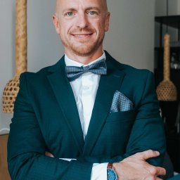 Decydujące Momenty Lidia Wawrzosek - Portret mężczyzny w ciemnozielonym garniturze, białej koszuli i muszce. Uśmiechnięty, z rękami skrzyżowanymi na piersiach. W tle widoczne elementy dekoracyjne.