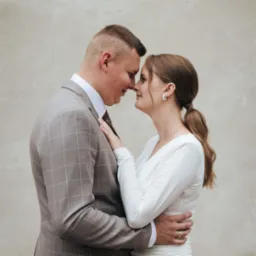 Zakochana para stoi blisko, dotykając się nosami. Mężczyzna w garniturze w kratę, kobieta w białej sukience. Delikatne oświetlenie, neutralne tło.