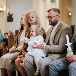 Decydujące Momenty Lidia Wawrzosek - Uroczystość chrztu: Dziewczynka w białej sukience siedzi na kolanach ojca w kościele, obok matka z kręconymi włosami i goście w ławkach. W rękach świeca.