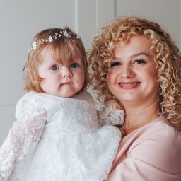 Decydujące Momenty Lidia Wawrzosek - Uśmiechnięta kobieta z kręconymi włosami trzyma na rękach niemowlę w białej sukience z koronką i ozdobą na głowie, na tle białych drzwi.