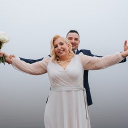 Uśmiechnięta para młoda w objęciach, ona trzyma bukiet białych róż, on obejmuje ją od tyłu. Mglista sceneria, woda w tle. Radosne uniesienie.