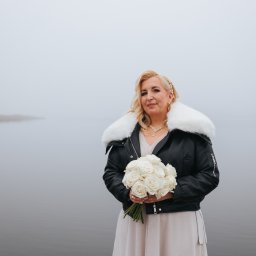 Panna młoda w skórzanej kurtce z futrzanym kołnierzem trzyma bukiet białych róż na tle mglistego jeziora. Nietypowa stylizacja ślubna.