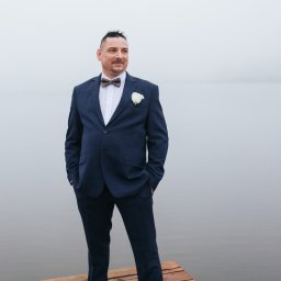 Elegancki mężczyzna w granatowym garniturze i muszce, z butonierką, stoi na drewnianym pomoście nad wodą w mglisty dzień, spoglądając w dal.