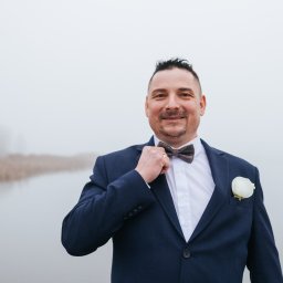 Uśmiechnięty mężczyzna w granatowym garniturze z butonierką i muszką, poprawiający ją na tle mglistego krajobrazu.