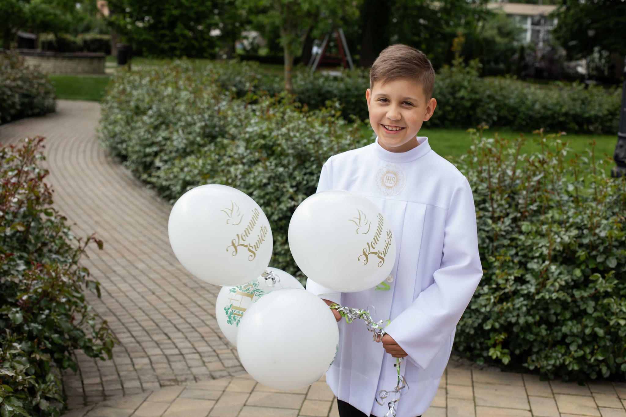 Uśmiechnięty chłopiec w albie komunijnej trzymający białe balony z napisem 'Komunia Święta' na tle parkowej alejki z zielenią.