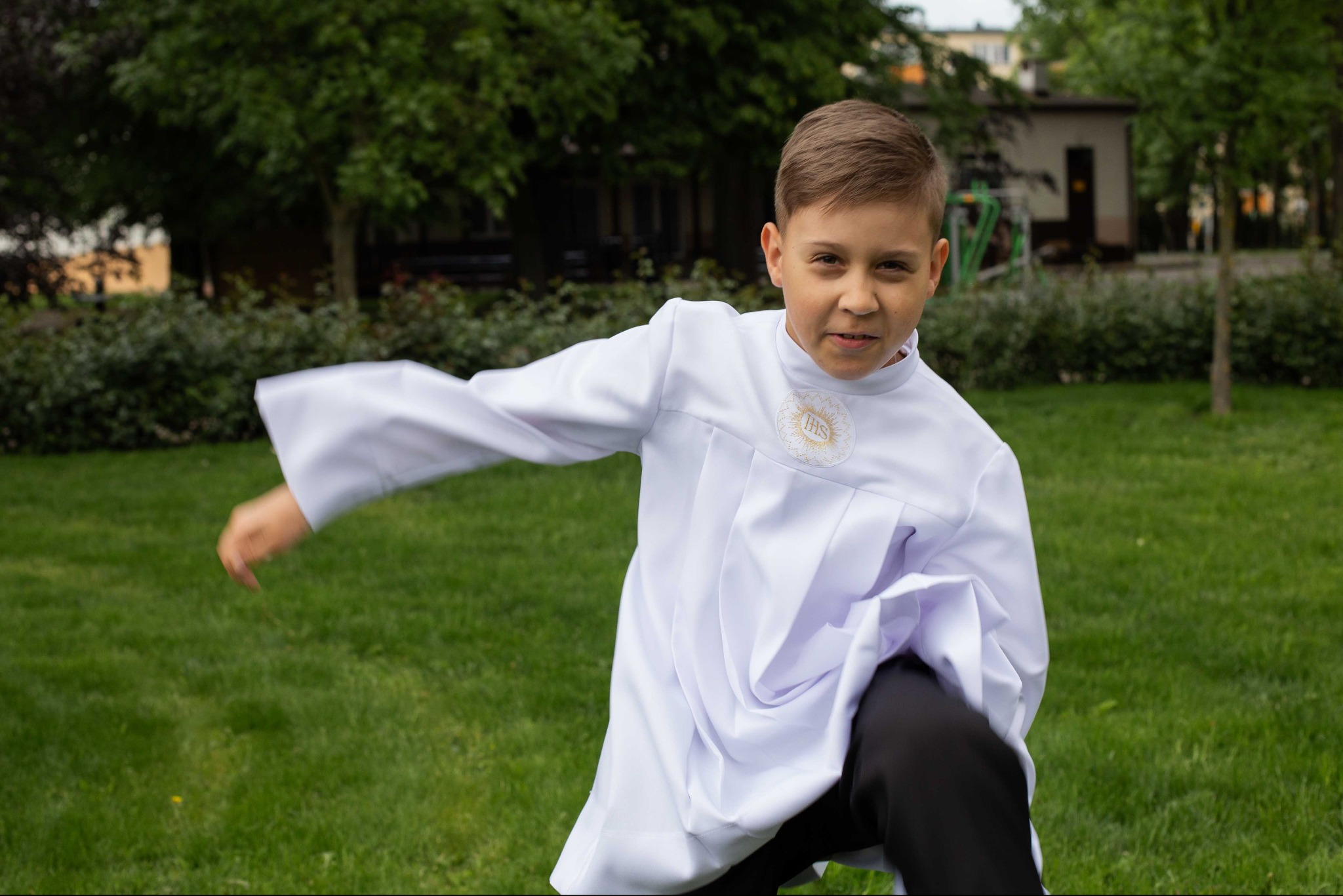 Uśmiechnięty chłopiec w białej albie komunijnej, w dynamicznej pozie na tle zielonego trawnika i drzew. Fotografia plenerowa z uroczystości Pierwszej Komunii Świętej.