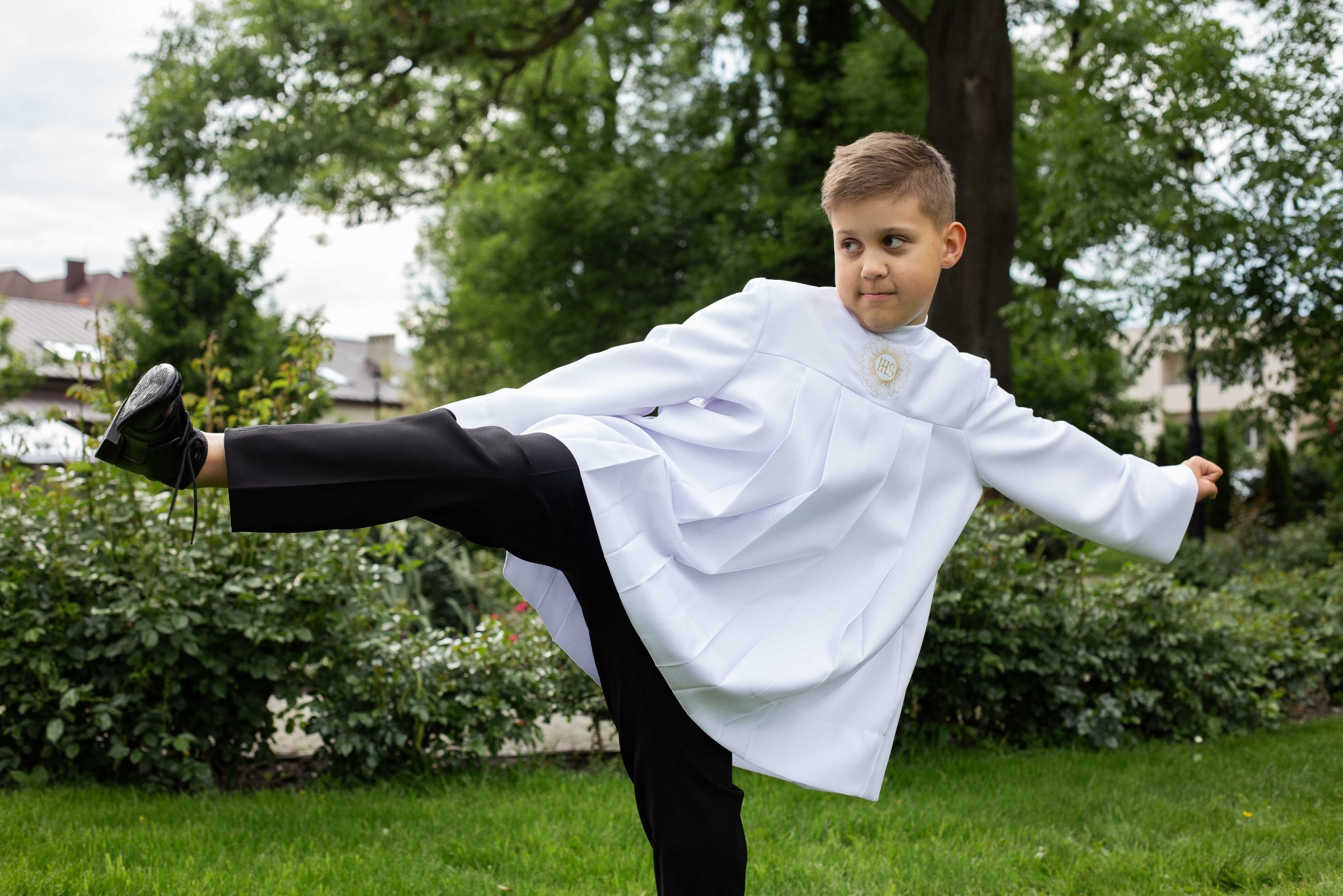 Chłopiec w stroju komunijnym ćwiczy równowagę na trawie, unosząc nogę. W tle zieleń drzew i krzewów, częściowo widoczny budynek. Ujęcie w plenerze, wesoła atmosfera.