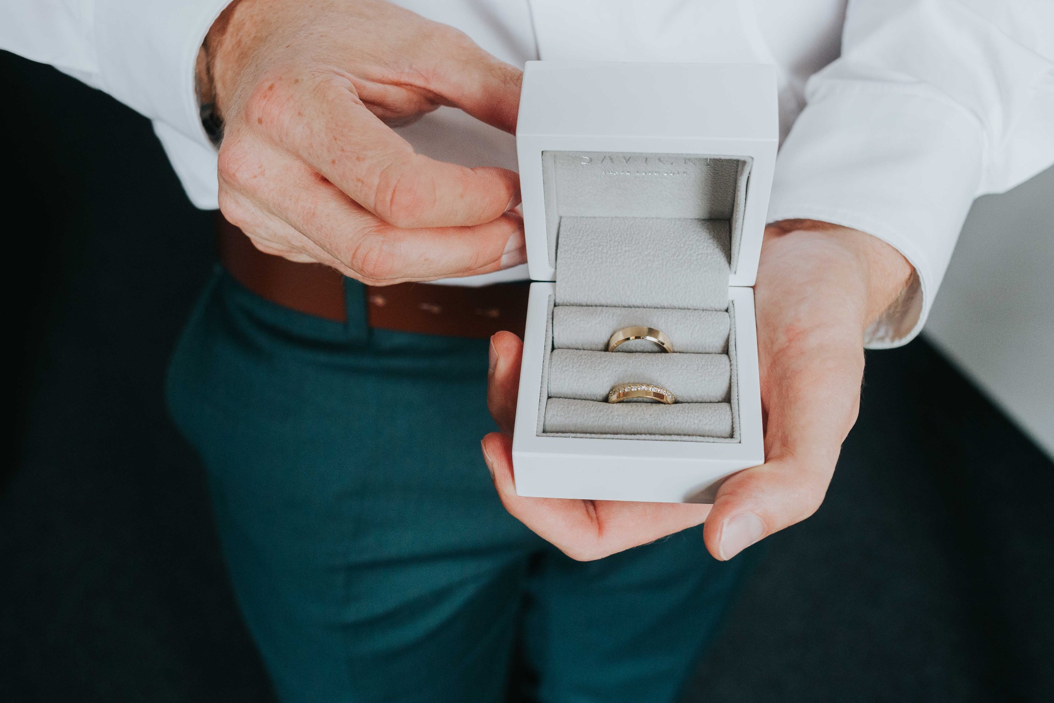 Dłonie trzymają białe pudełko z dwoma złotymi obrączkami. Obrączka z brylantami na dole. Mężczyzna ubrany w białą koszulę i zielone spodnie.