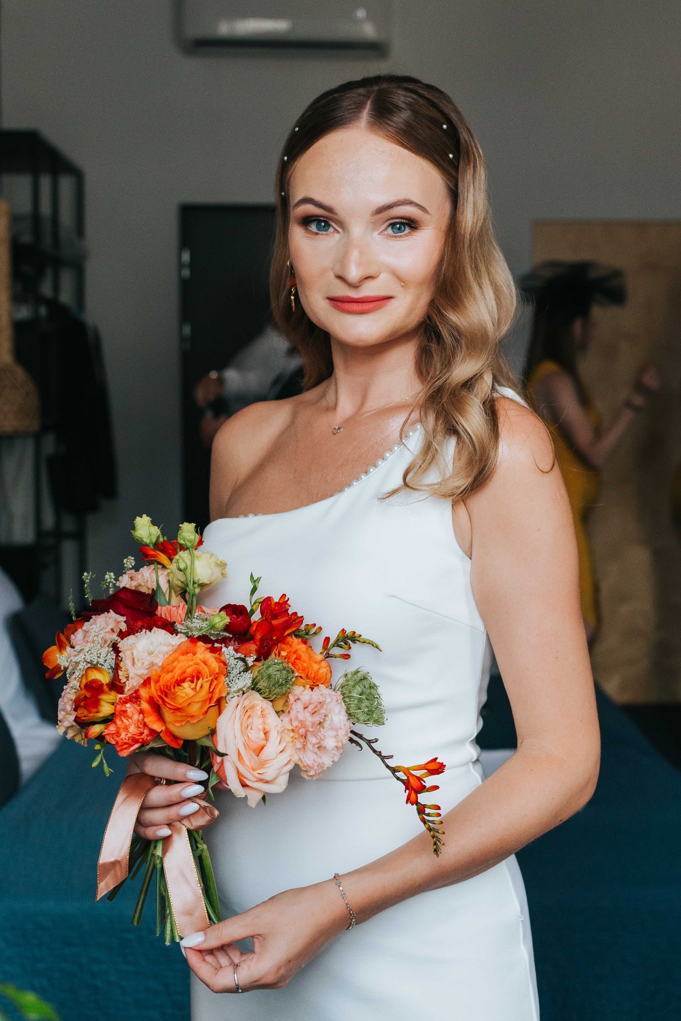 Panna młoda w białej sukni na jedno ramię trzyma bukiet z różowych i pomarańczowych kwiatów. Uśmiechnięta, z blond włosami i subtelnym makijażem.