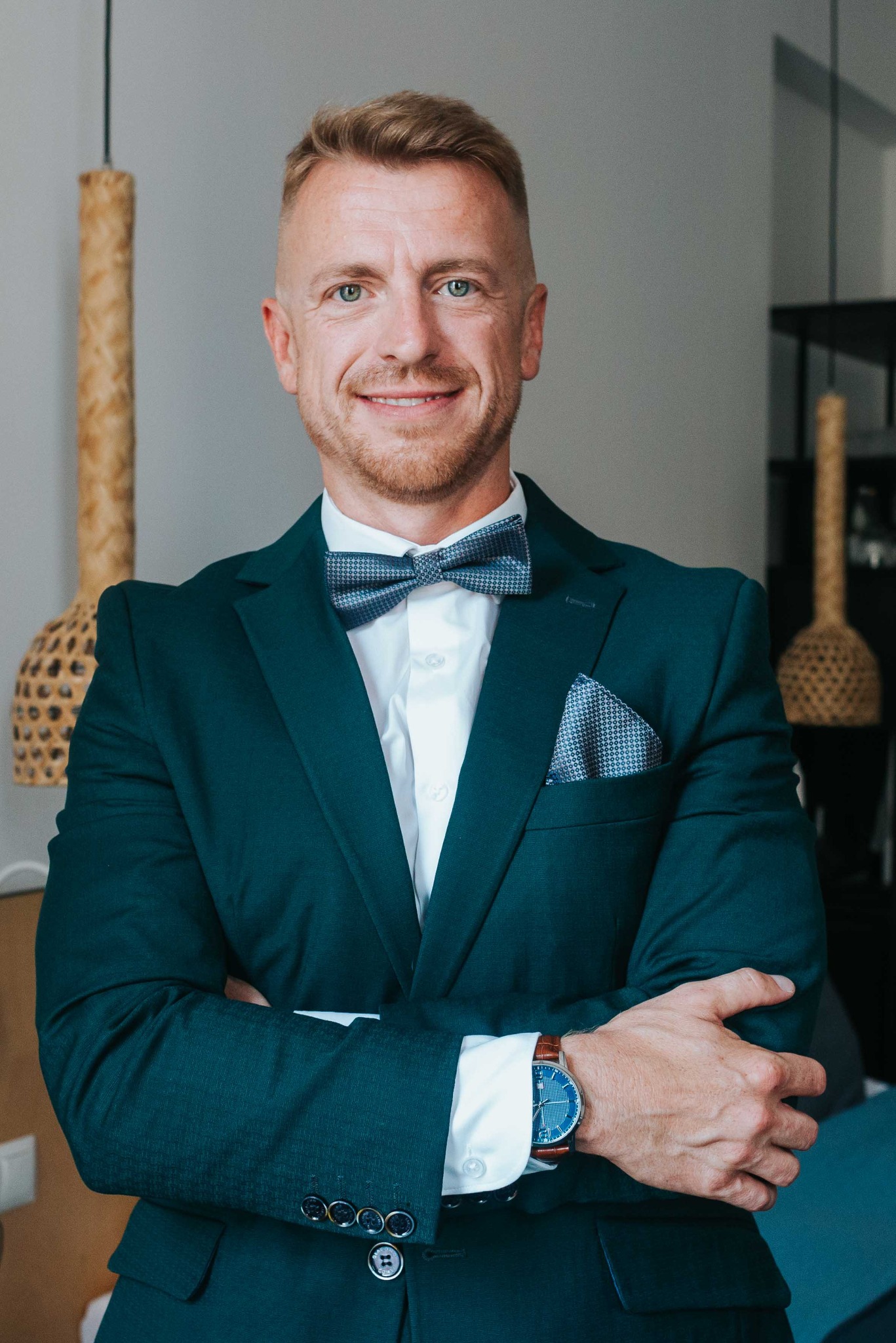 Portret mężczyzny w ciemnozielonym garniturze, białej koszuli i muszce. Uśmiechnięty, z rękami skrzyżowanymi na piersiach. W tle widoczne elementy dekoracyjne.
