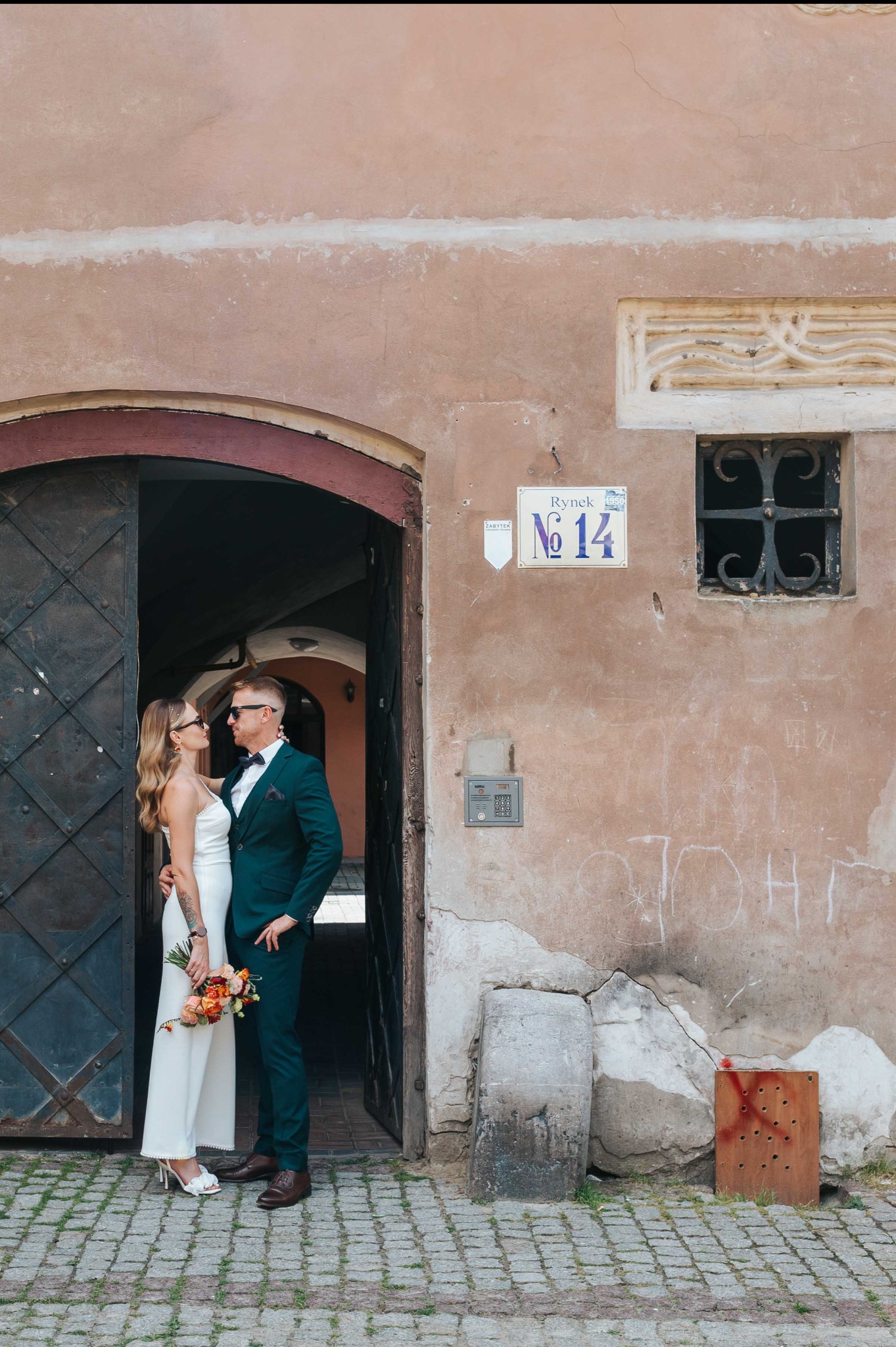 Para młoda w eleganckich strojach pozuje w bramie kamienicy z tabliczką 'Rynek No 14'. Ona w białym kombinezonie, on w zielonym garniturze. Uśmiechy, kwiaty.