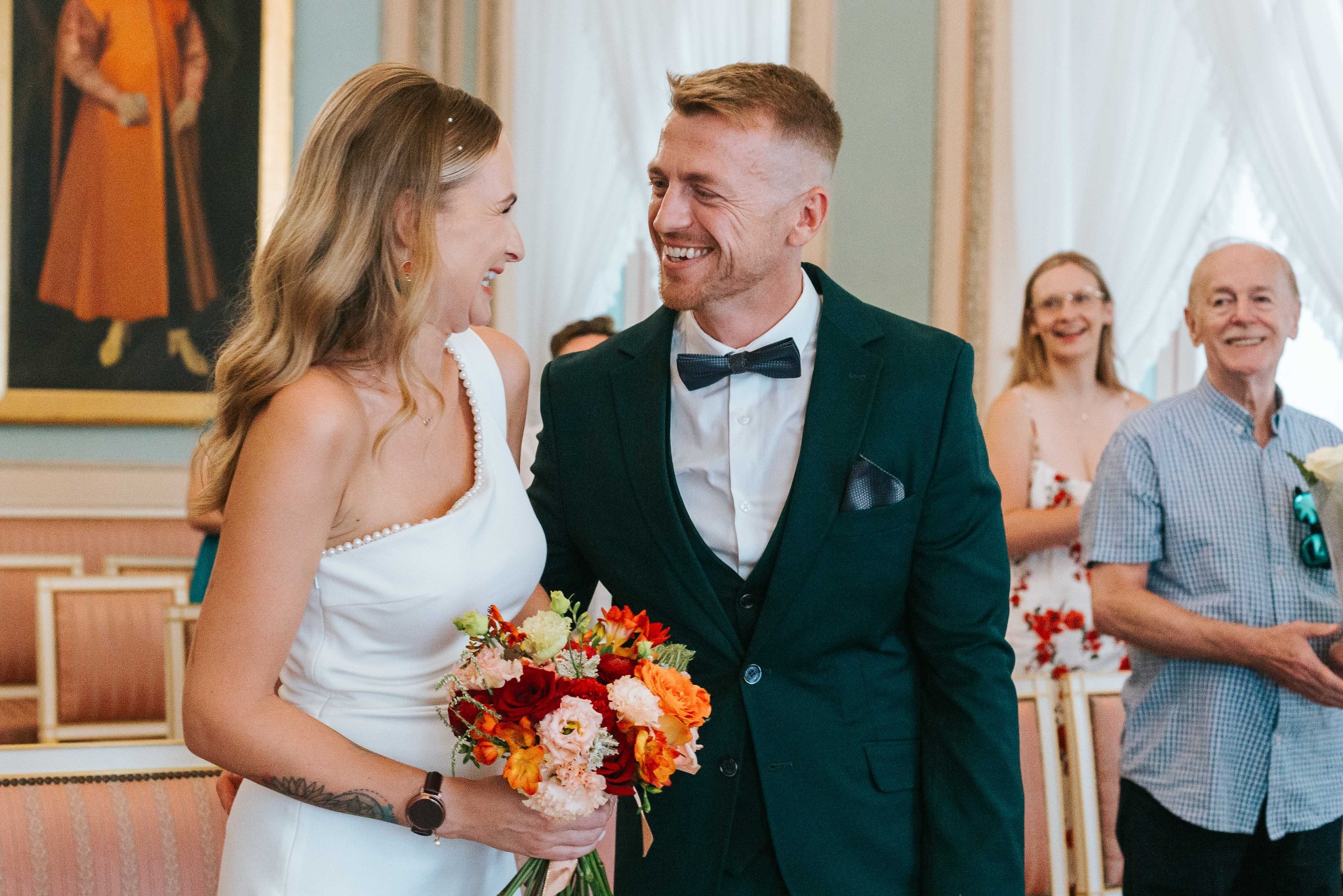 Uśmiechnięta para młoda w eleganckich strojach, trzymająca bukiet kwiatów, z gośćmi w tle podczas ceremonii ślubnej w jasnym wnętrzu.