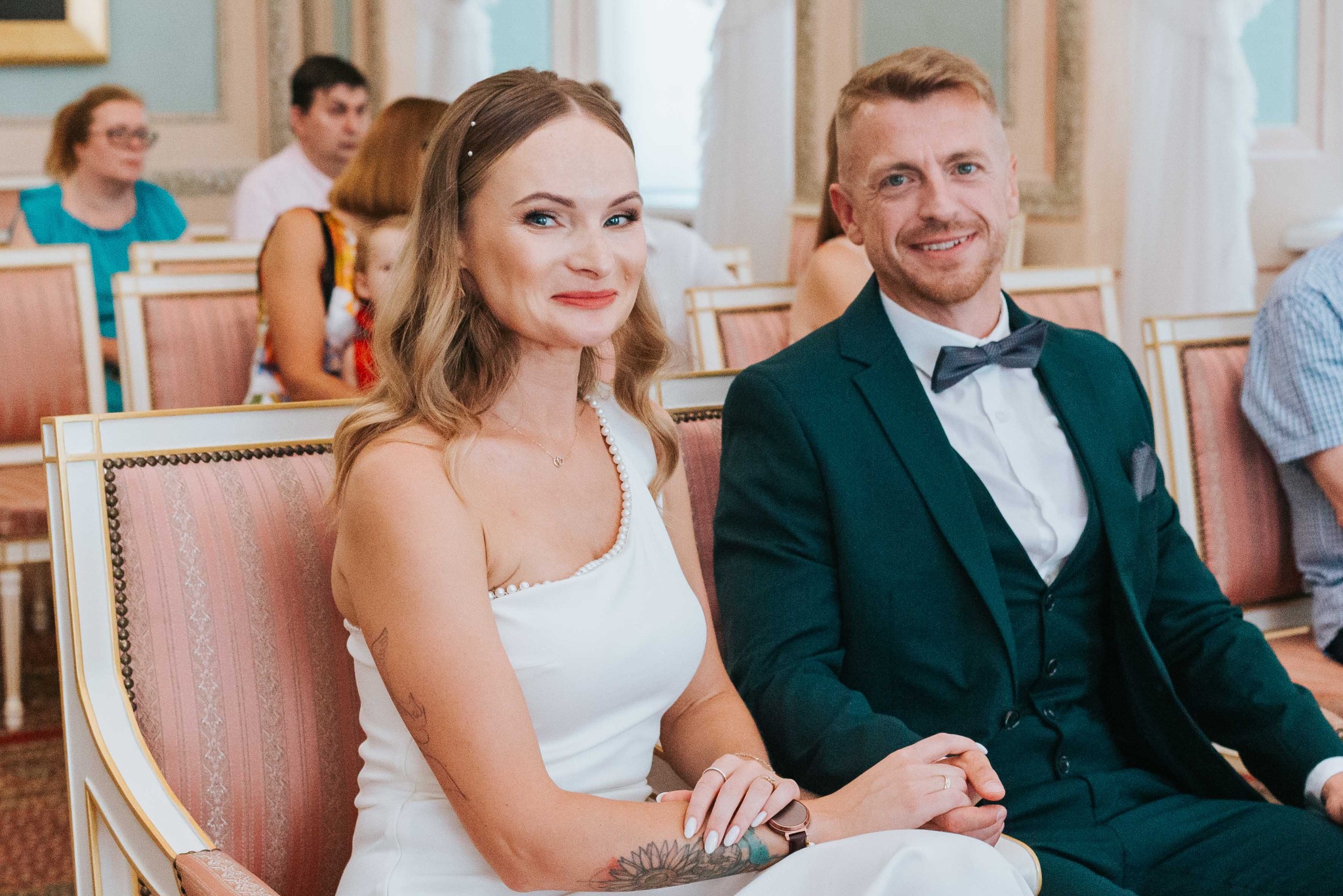 Elegancka para młoda siedzi obok siebie na tle gości weselnych. Panna młoda w białej sukni, pan młody w ciemnozielonym garniturze. Uśmiechy, ciepłe oświetlenie.