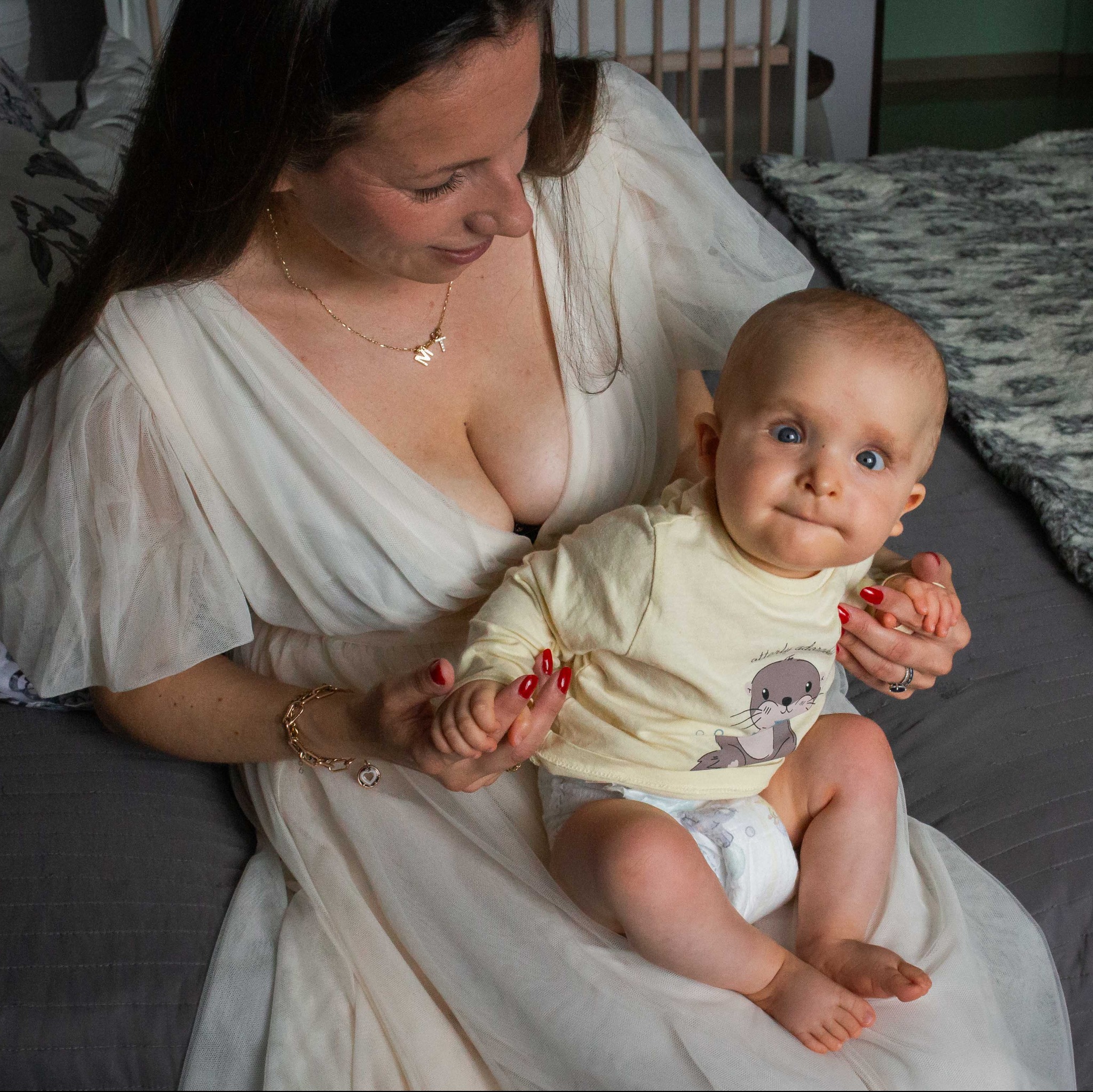 Mama w zwiewnej sukience trzyma na kolanach niemowlę w koszulce z wydrą, w tle łóżeczko dziecięce. Uśmiechnięta kobieta z biżuterią patrzy na dziecko.