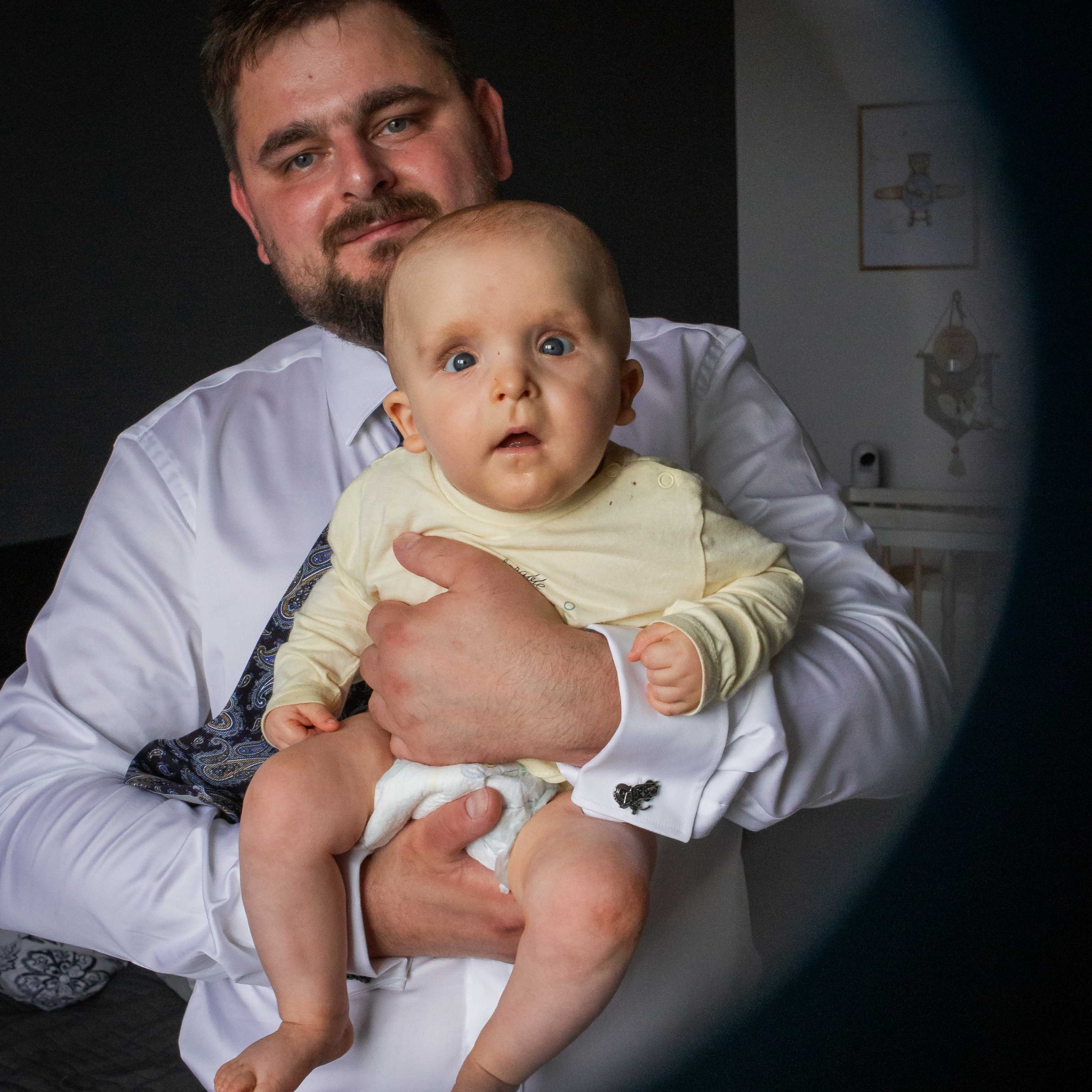 Tata w białej koszuli i mankietach z motywem gitary trzyma na rękach niemowlę w żółtym ubranku. W tle pokój dziecięcy z łóżeczkiem i rysunkiem samolotu.