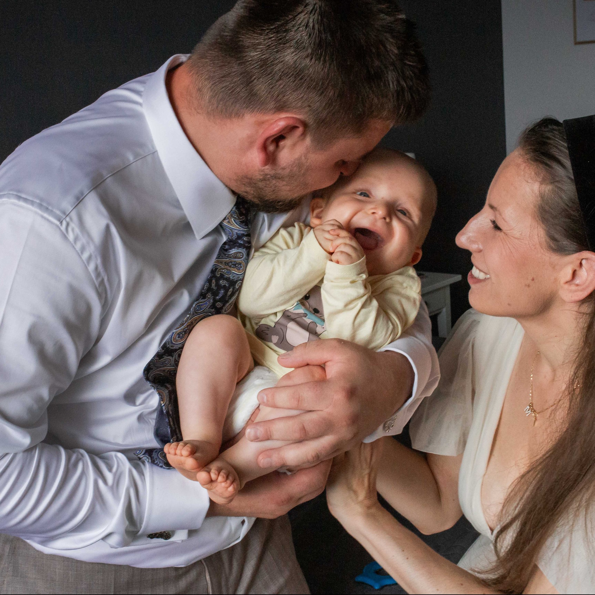 Uśmiechnięte niemowlę na rękach taty, który całuje je w głowę. Mama patrzy z uśmiechem. Zdjęcie rodzinne w jasnych barwach na ciemnym tle.