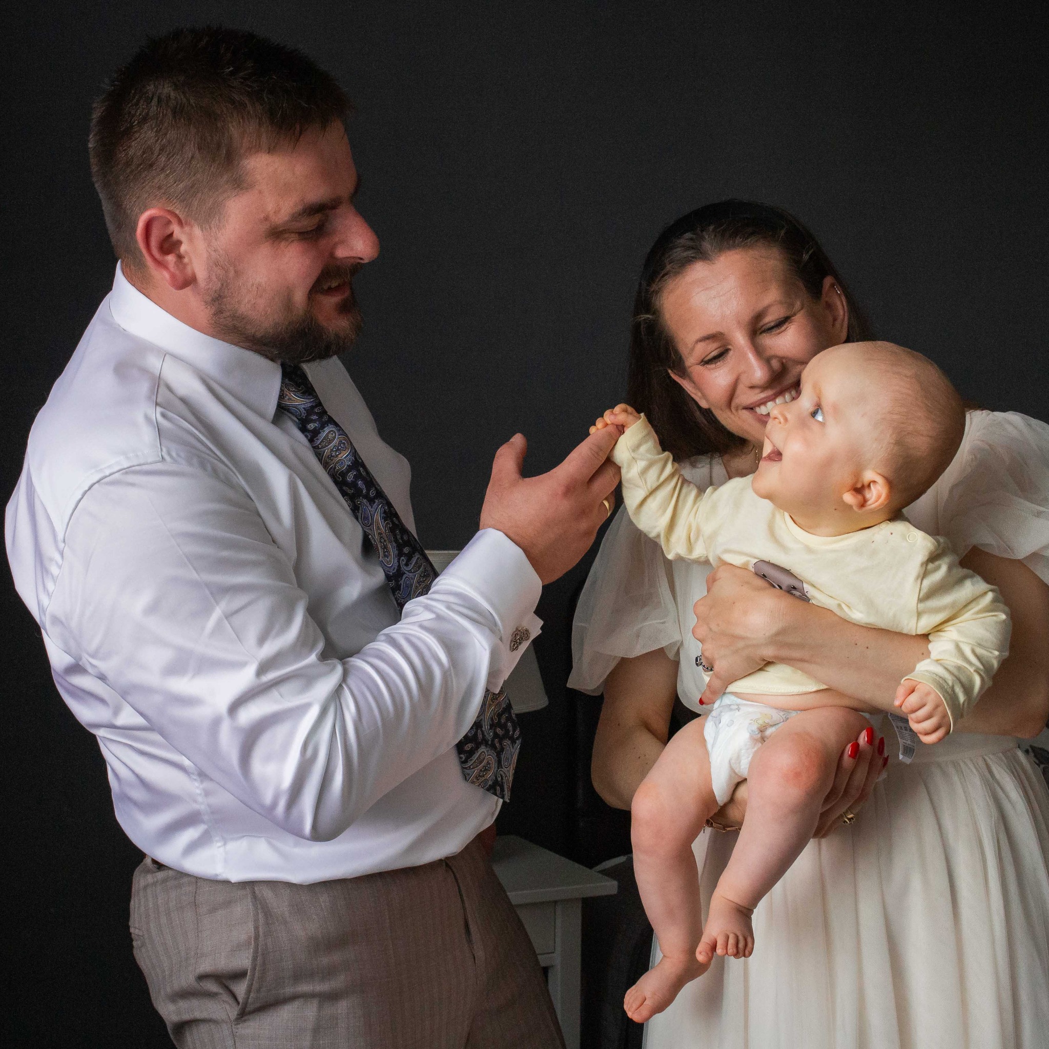 Rodzinne ujęcie w studio: uśmiechnięta matka trzyma niemowlę, które wyciąga rączkę do ojca w koszuli i krawacie. Szczęśliwa, ciepła atmosfera.