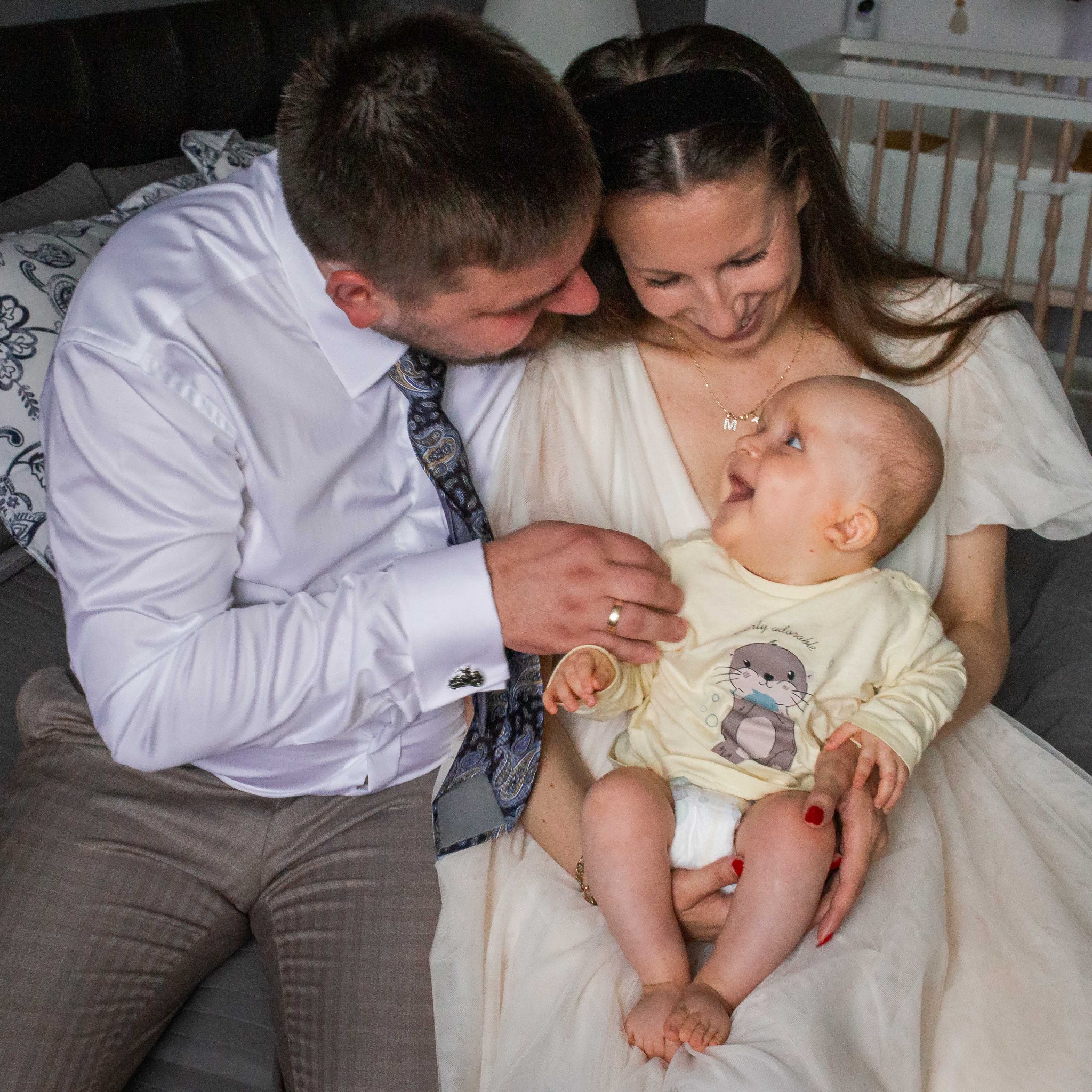 Szczęśliwa rodzina: mama w kremowej sukience trzyma uśmiechnięte niemowlę, tata w koszuli i krawacie patrzy z czułością. W tle łóżeczko dziecięce.