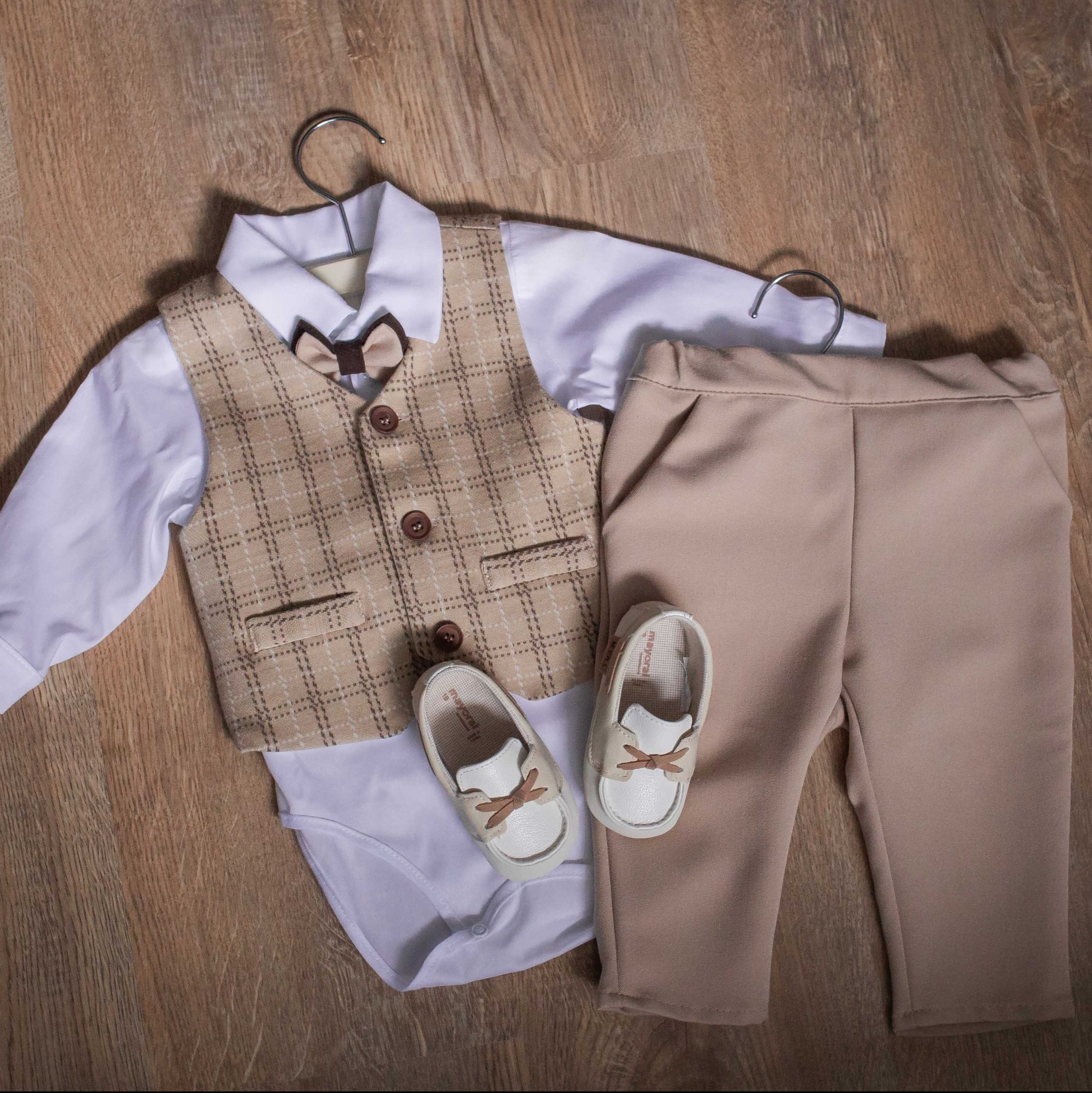 Elegancki komplet niemowlęcy: body z muchą, kamizelka w kratę, beżowe spodnie i buciki na drewnianej podłodze. Ubranko zawieszone na wieszakach.
