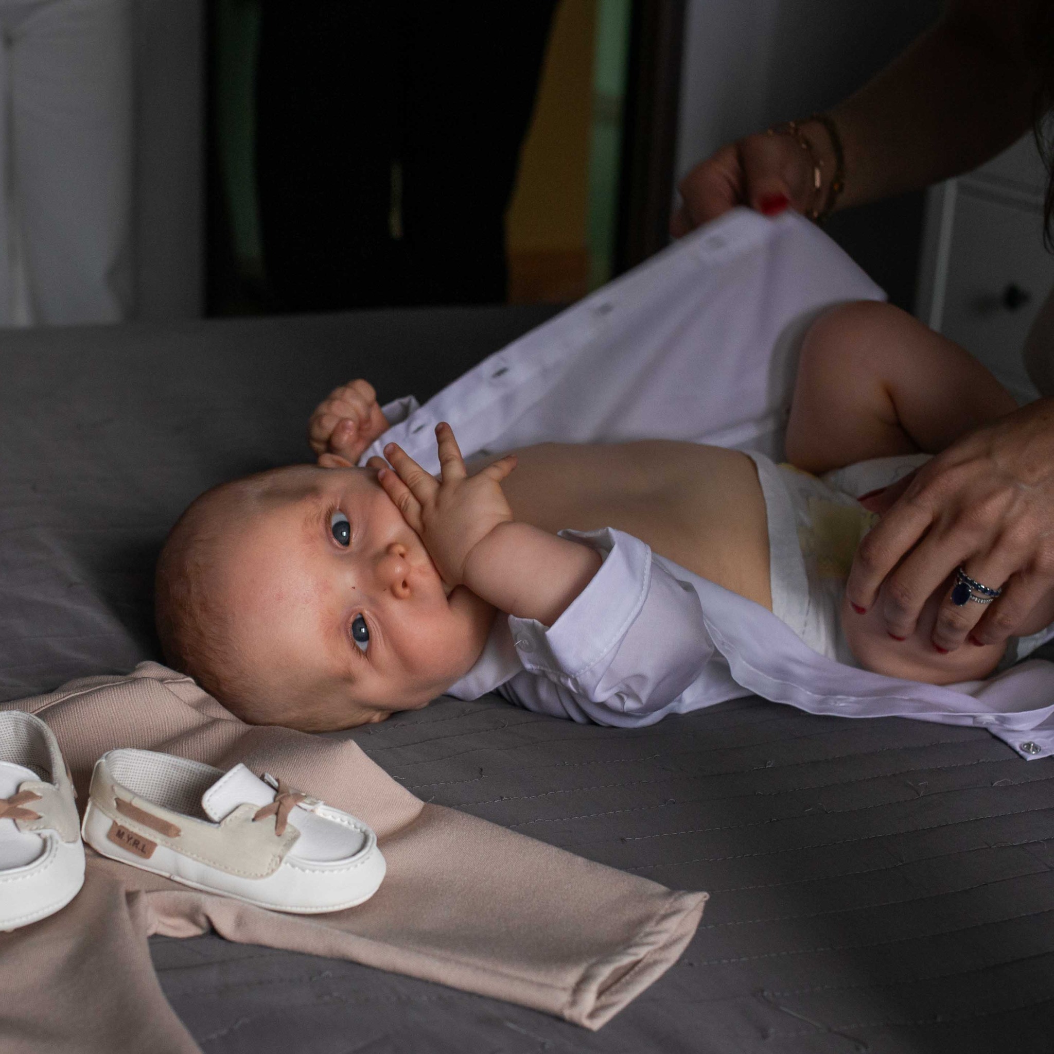 Urocze niemowlę leży na szarym kocyku, ubrane w białą koszulę, z parą małych, beżowo-białych bucików i beżowymi spodniami obok. Matka pomaga mu się ubrać.
