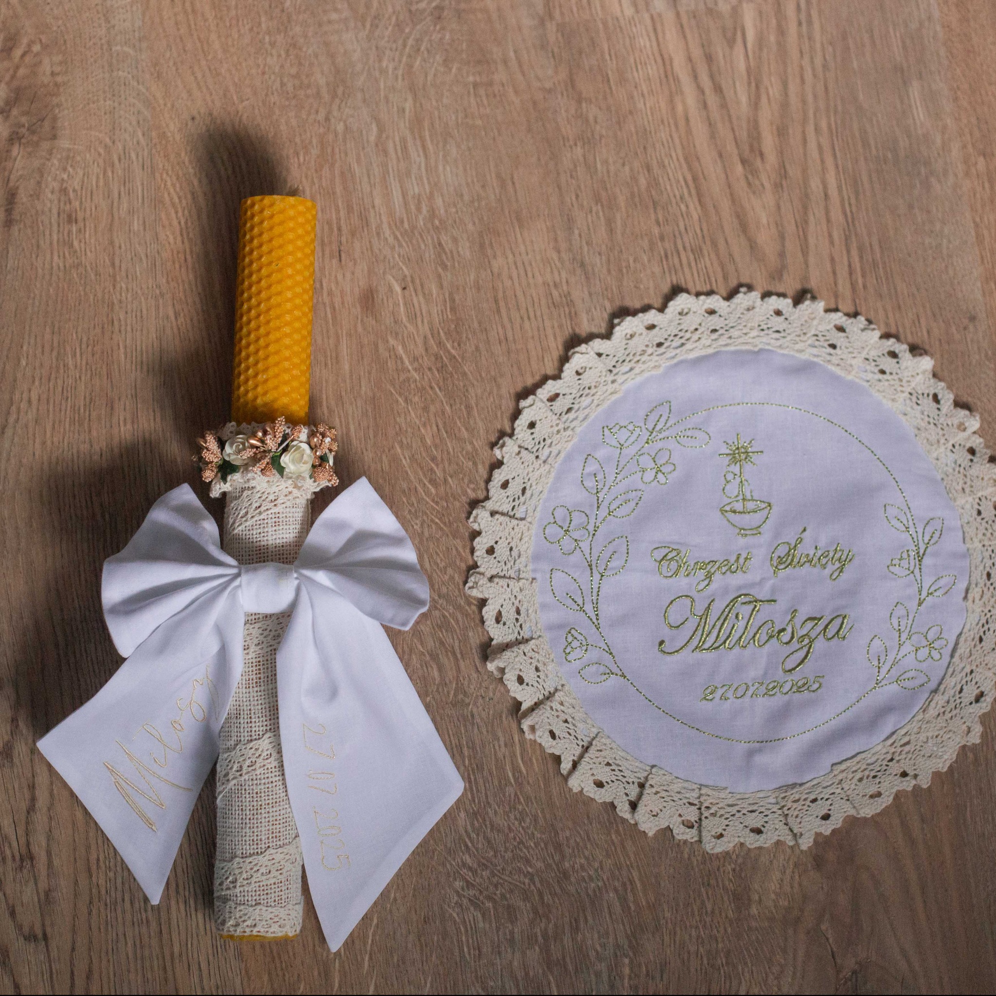 Dekoracyjna świeca z kokardą i haftowana serwetka pamiątka Chrztu Świętego Miłosza z datą 27.07.2025, leżące na drewnianym stole.