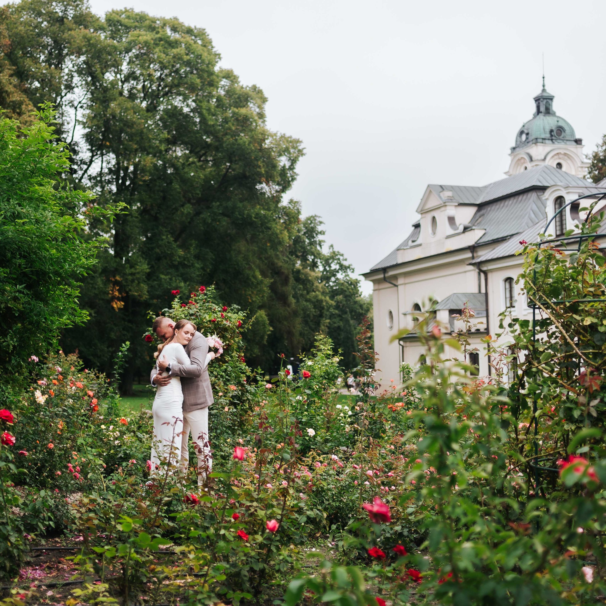 Para młoda w objęciach wśród róż w ogrodzie w Lublinie. W tle budynek z kopułą. Romantyczna sceneria i naturalne światło. Fotografia ślubna.