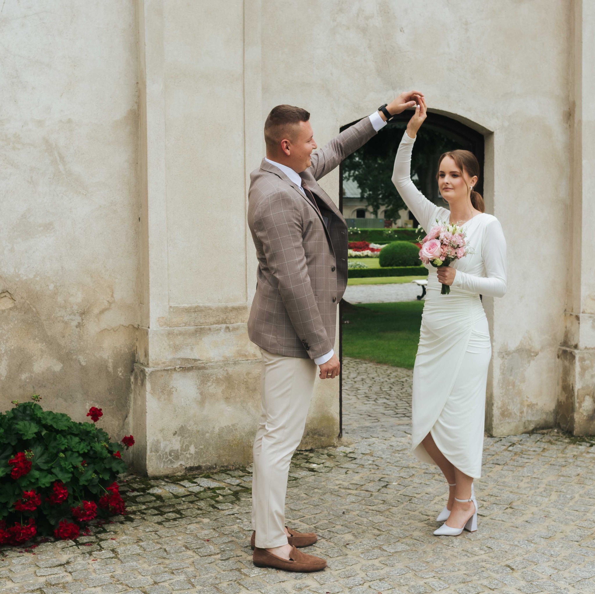 Para młoda tańczy w otoczeniu architektury. Panna młoda w białej sukni trzyma bukiet różowych kwiatów. Pan młody w brązowym garniturze. Romantyczna sceneria.