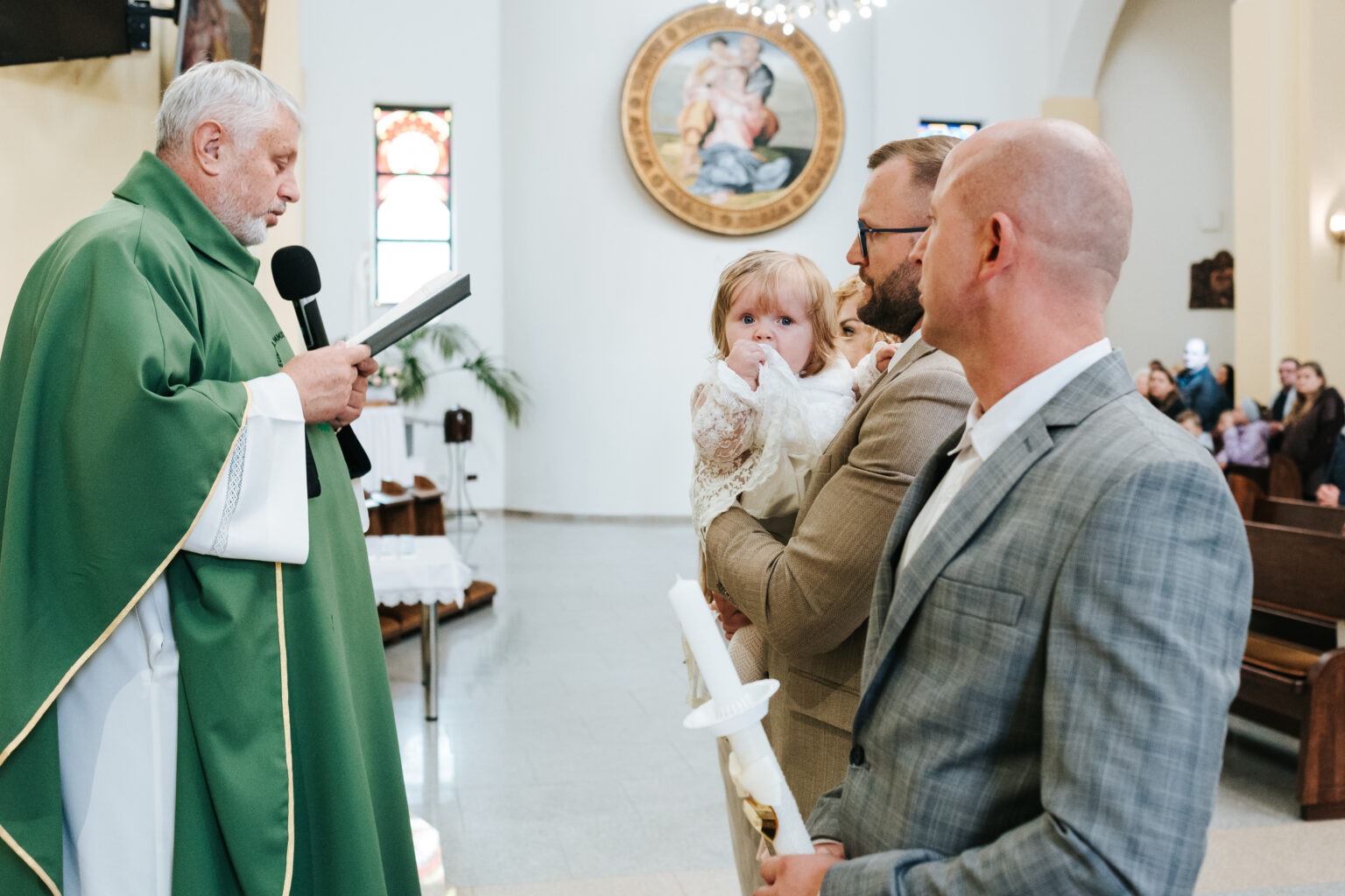 Ksiądz w zielonej szacie prowadzi ceremonię chrztu w kościele. Rodzice trzymają dziecko ubrane w białą szatkę, w tle widoczni goście.