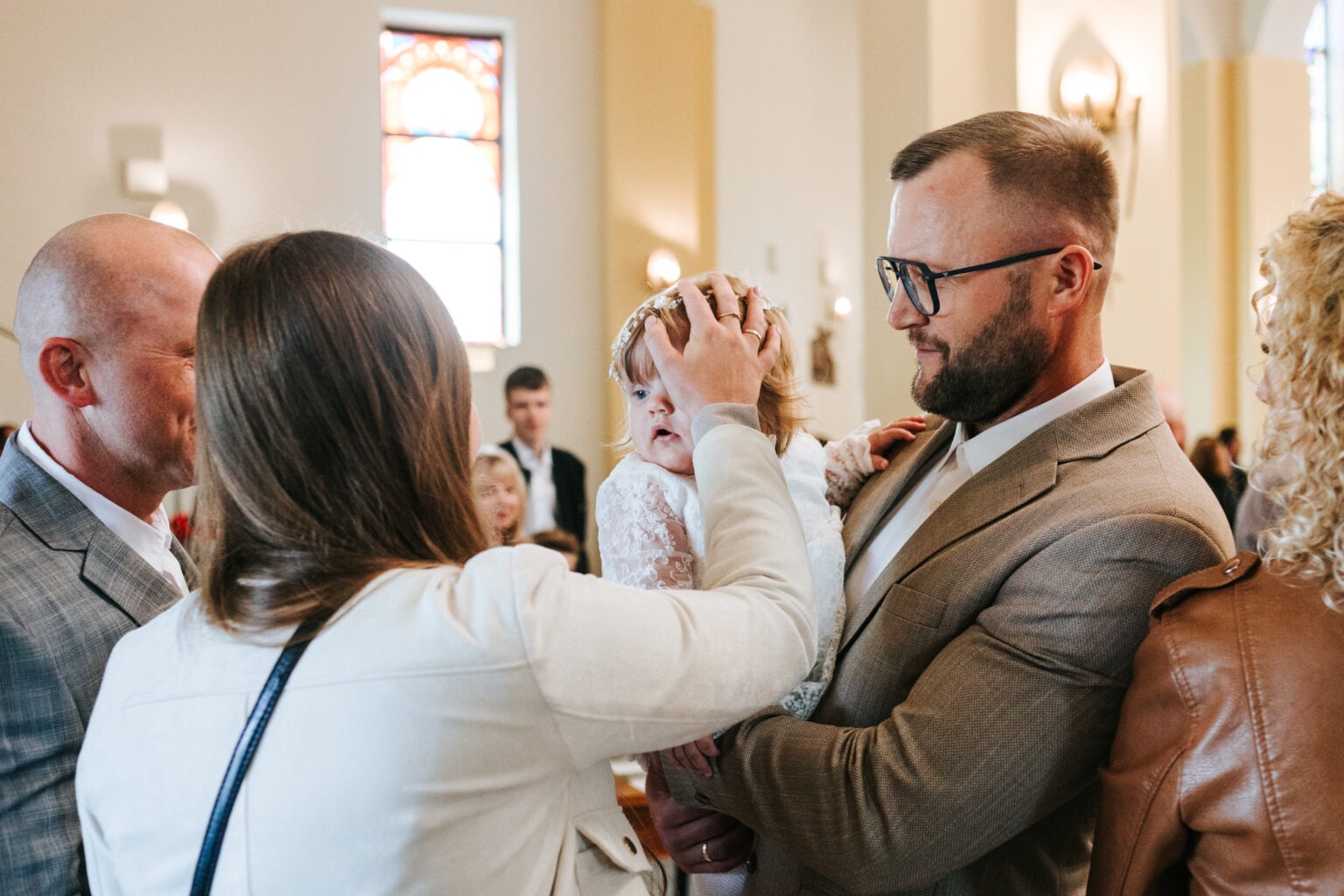 Uroczystość chrztu: ojciec trzyma niemowlę w białej sukience, matka poprawia dziecku wianek na głowie, w tle goście w kościele.