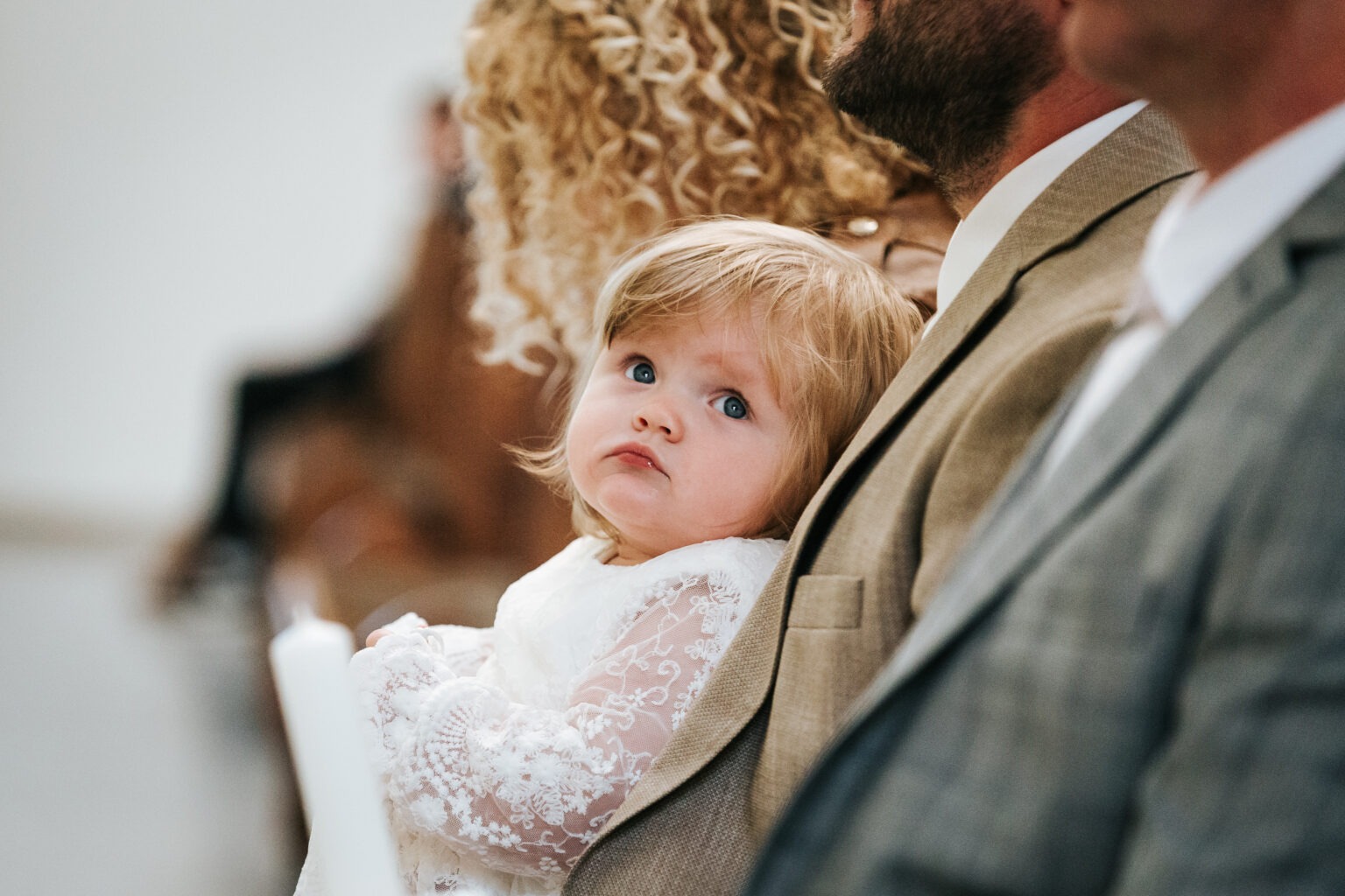 Dziewczynka w białej, koronkowej sukience, zamyślona, oparta o ramię mężczyzny w garniturze. W tle widoczna osoba z kręconymi włosami.