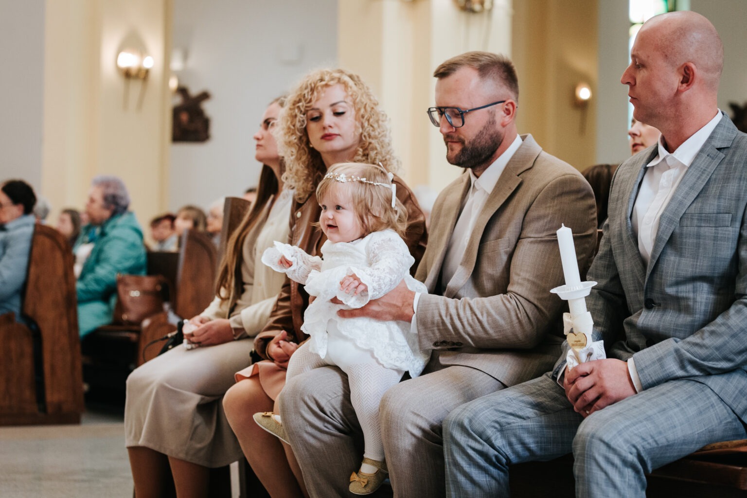 Uroczystość chrztu: Dziewczynka w białej sukience siedzi na kolanach ojca w kościele, obok matka z kręconymi włosami i goście w ławkach. W rękach świeca.
