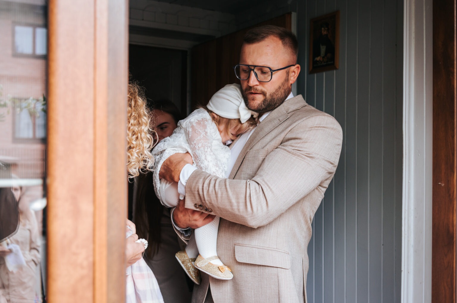 Elegancki mężczyzna w beżowym garniturze trzyma śpiące dziecko w białej sukience i czapeczce, w tle widoczni inni ludzie i fragment wnętrza.