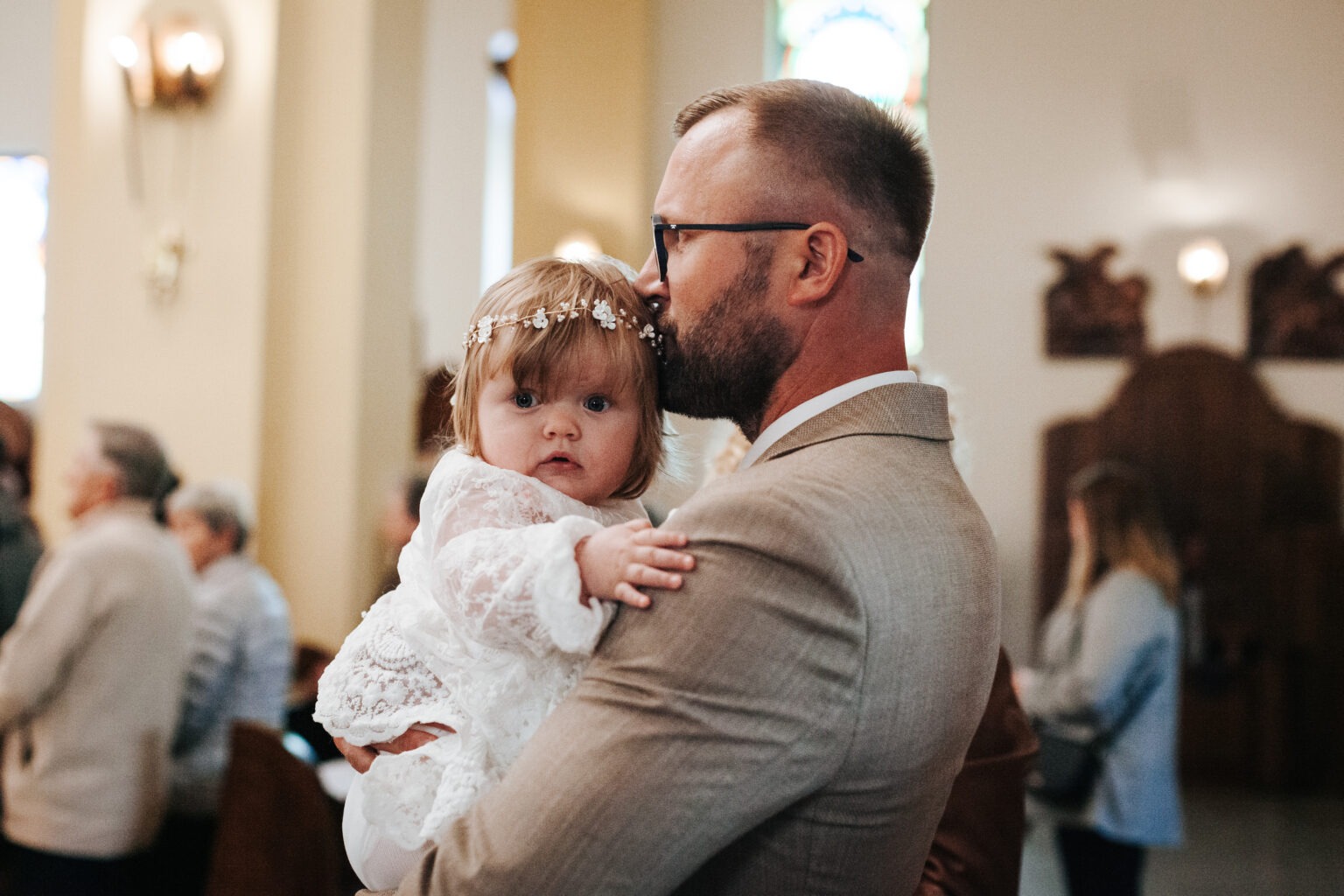 Ojciec w beżowym garniturze całuje w czoło uroczą dziewczynkę w białej koronkowej sukience i wianku na głowie, w kościele podczas uroczystości.
