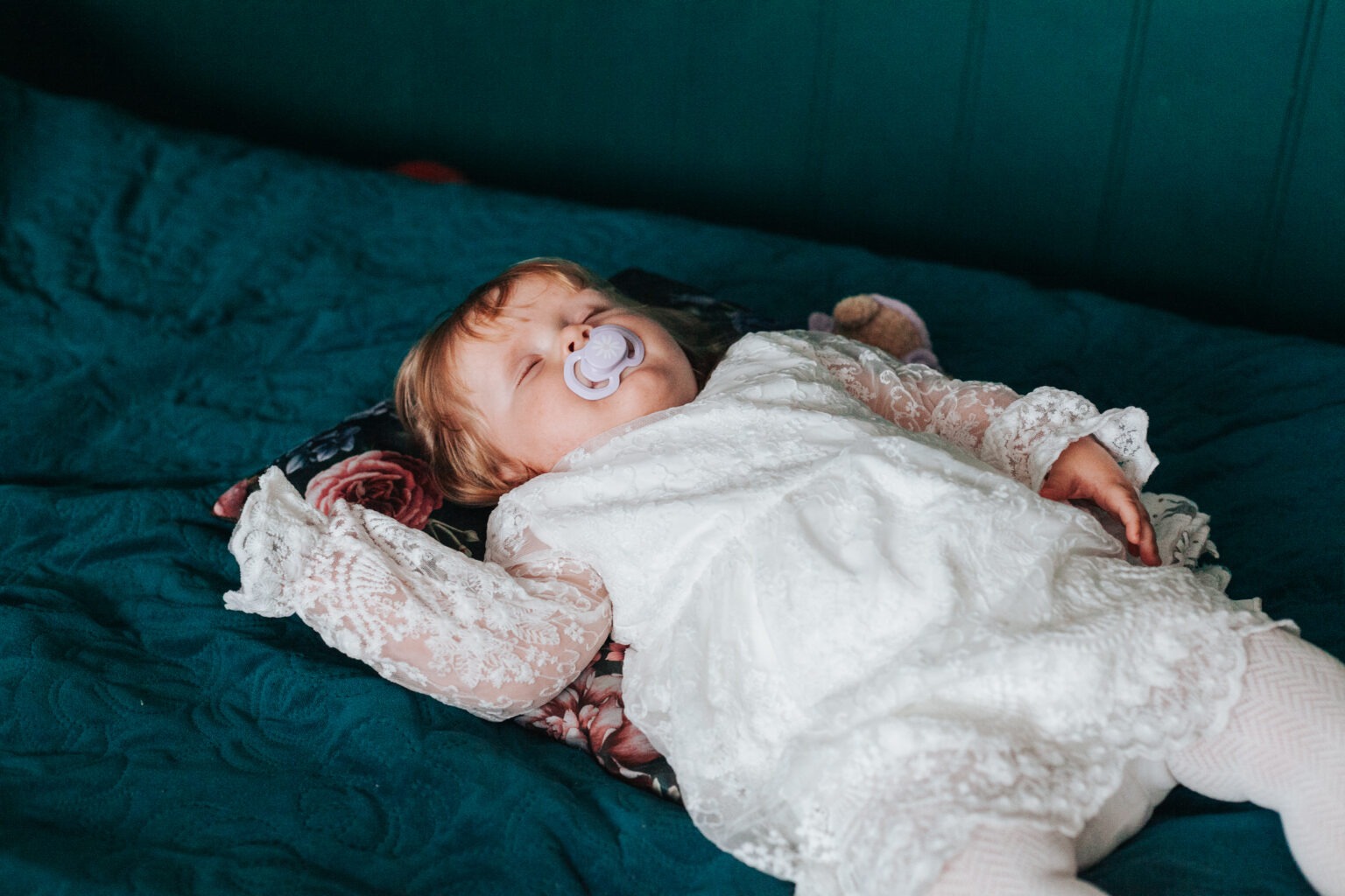 Śpiąca dziewczynka w białej, koronkowej sukience leży na poduszce z motywem kwiatowym na tle turkusowej narzuty. Dziecko ma smoczek w ustach.