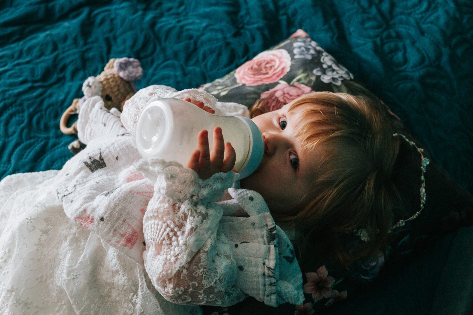 Dziewczynka z rudymi włosami pije z butelki leżąc na wzorzystej poduszce i turkusowym kocu. Ubrana w białą koronkową sukienkę, obok pluszowy miś.