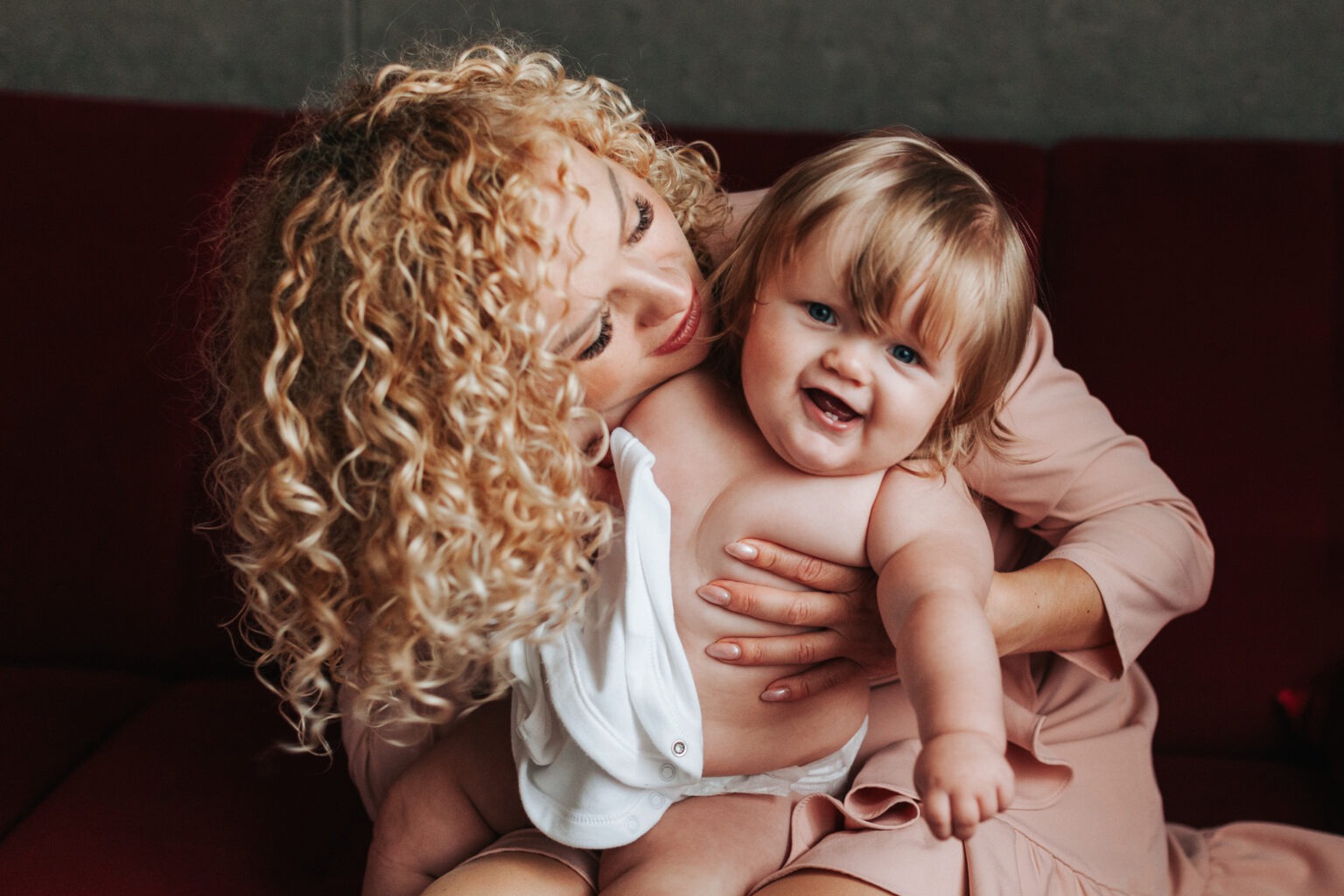 Uśmiechnięta blondynka z burzą loków tuli roześmianego niemowlaka na tle bordowej sofy. Delikatne, naturalne światło podkreśla bliskość i radość.