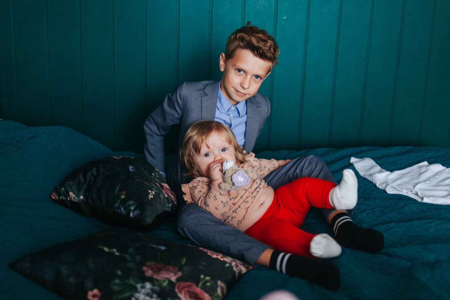 Portret rodzeństwa na łóżku: starszy brat w garniturze i młodsza siostra z pluszakiem na tle turkusowej ściany. Ujęcie z bliska, naturalne światło.