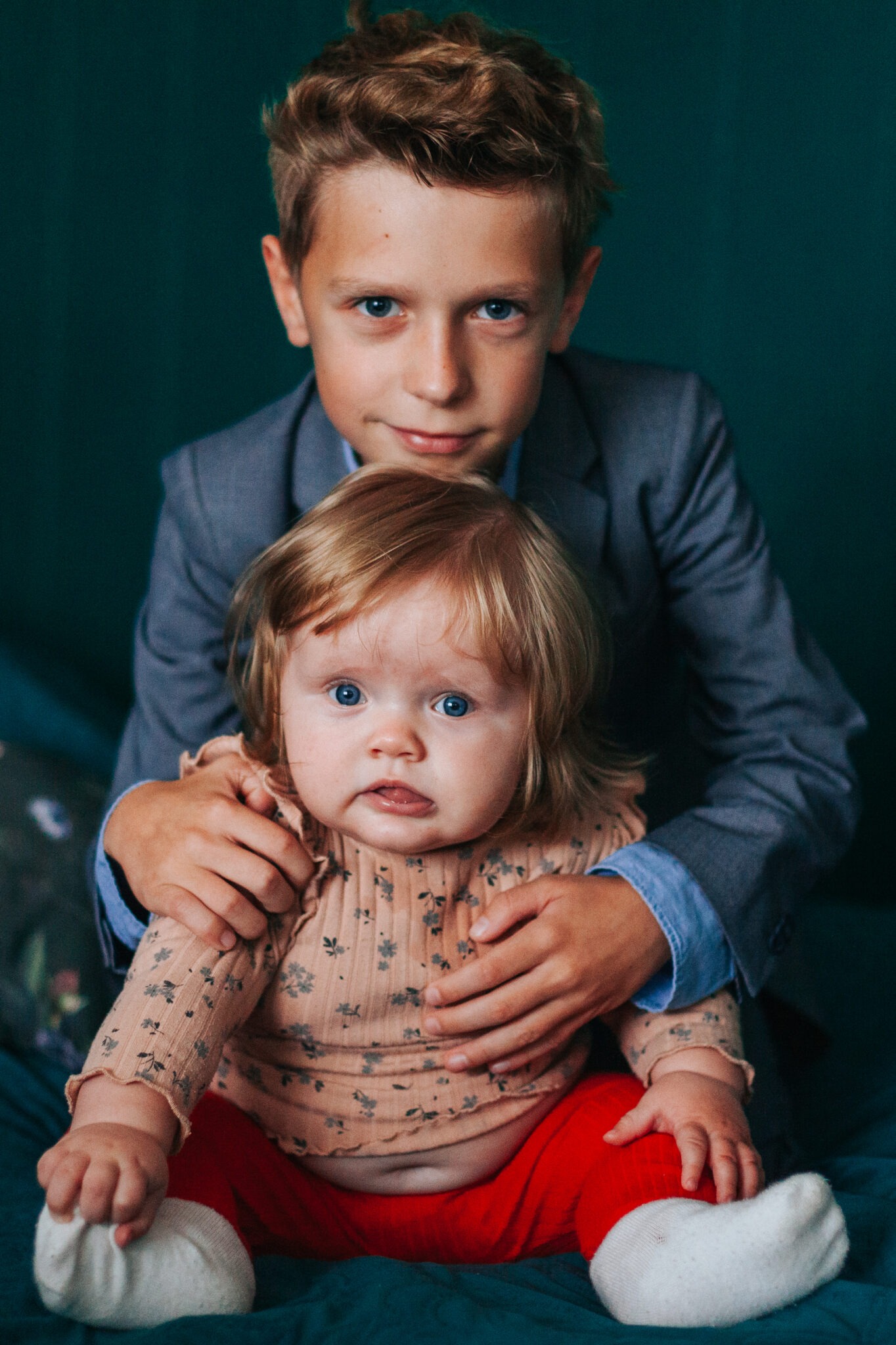Portret rodzeństwa: starszy brat w szarej marynarce obejmuje młodszą siostrę w czerwonych spodniach na tle ciemnozielonej ściany. Dzieci patrzą w obiektyw.