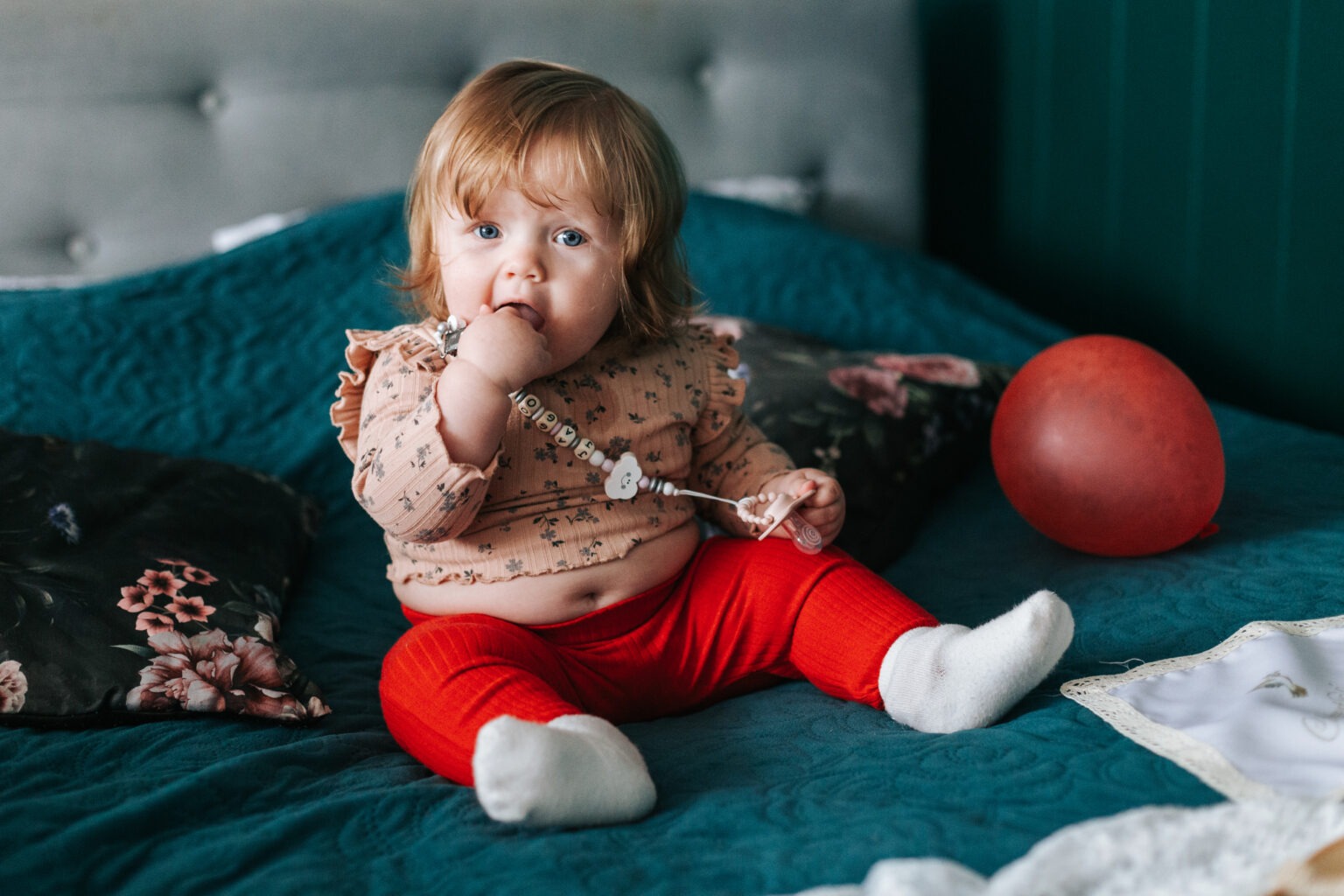 Siedzące na turkusowym łóżku niemowlę w czerwonych spodniach i bluzce w kwiatki, z imiennym smoczkiem w buzi, obok czerwony balon i poduszka w kwiaty.