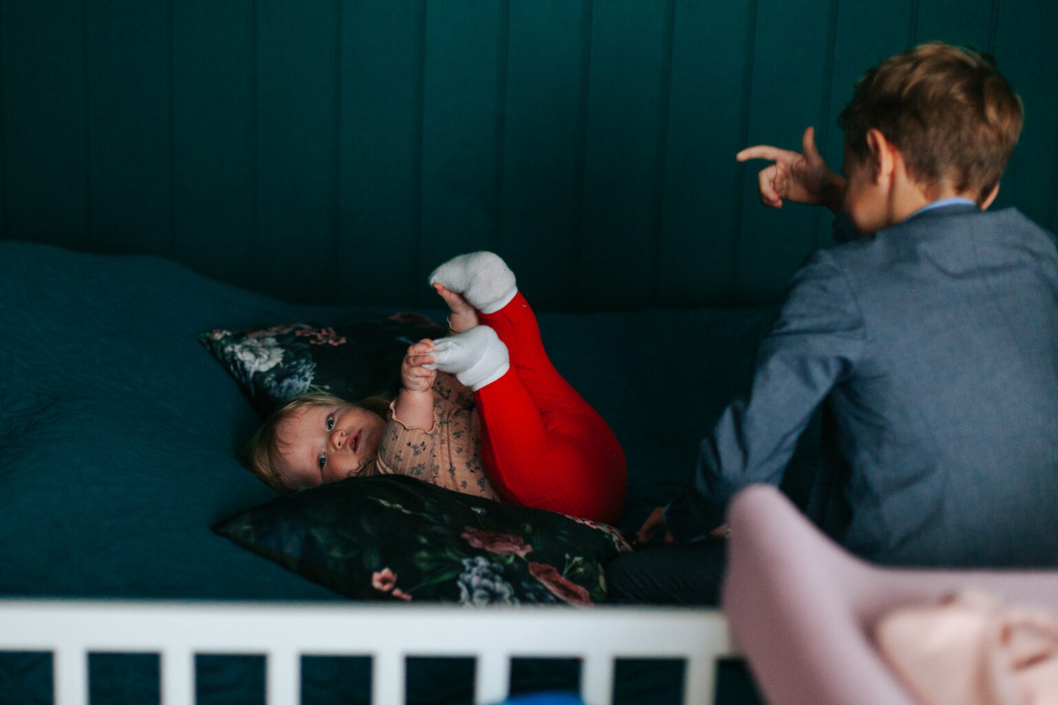 Uśmiechnięta niemowlęca dziewczynka w czerwonych legginsach leży na łóżku, trzymając stopy. Chłopiec w marynarce wskazuje palcem w jej stronę na tle turkusowej ściany.