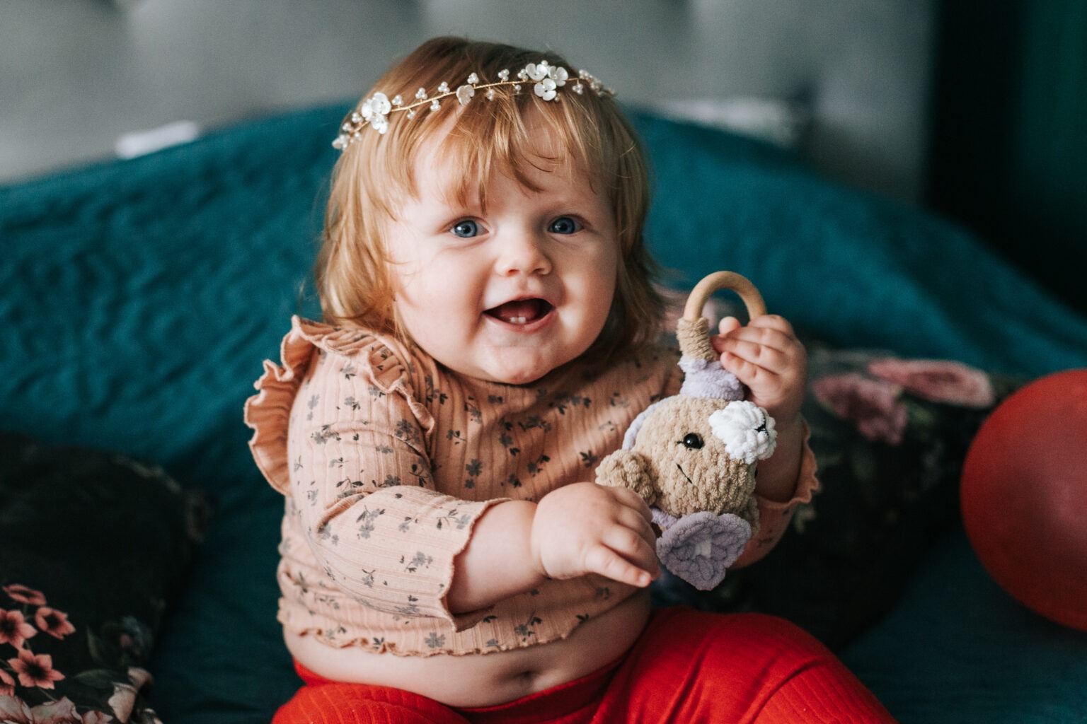 Uśmiechnięta blond dziewczynka w wianku trzyma pluszowego króliczka na tle turkusowej kanapy i czerwonego balona. Dziecko w bluzce w kwiatki i czerwonych spodniach.