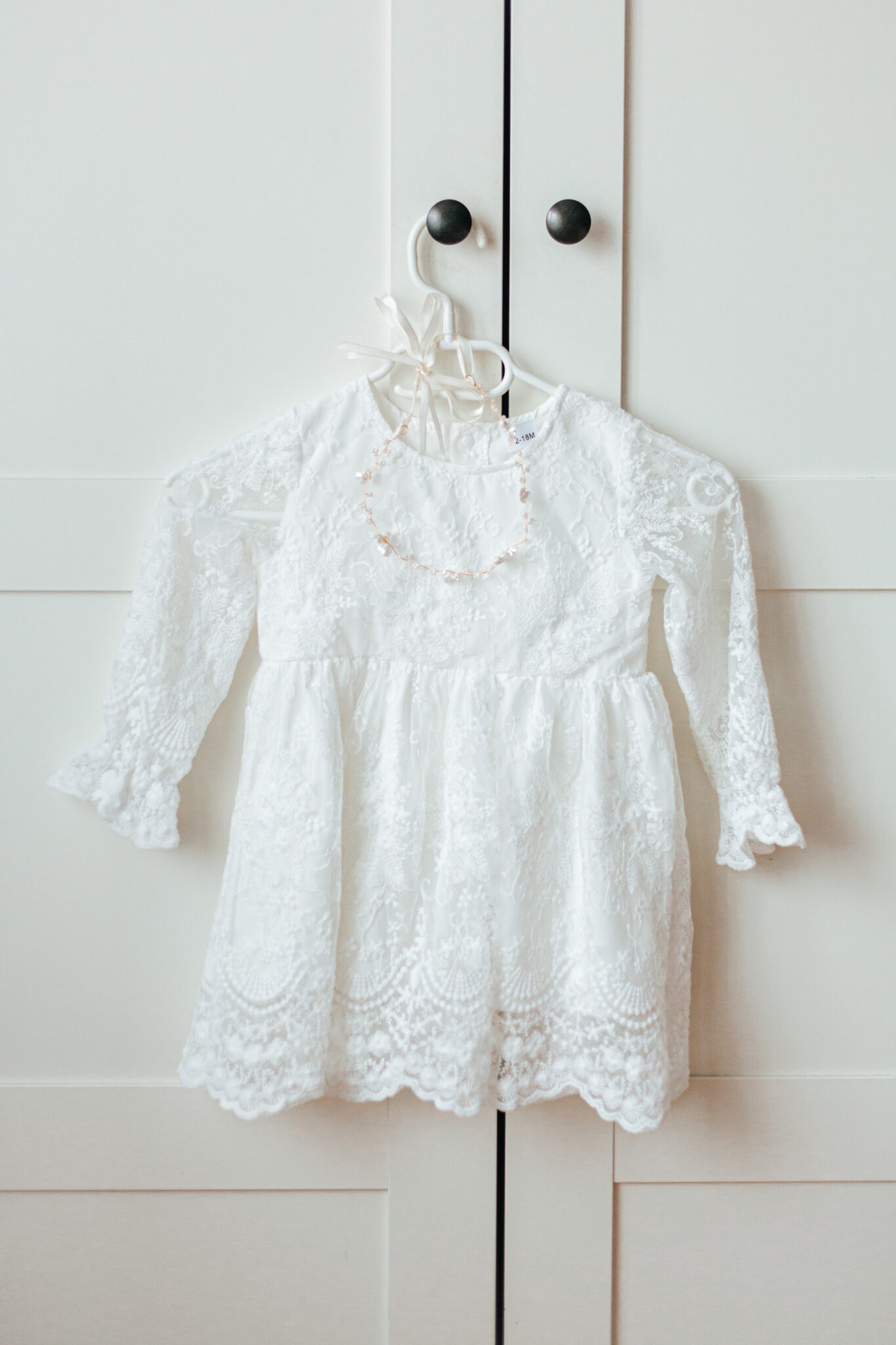 Koronkowa sukienka dziewczęca w kolorze ecru, zawieszona na białym wieszaku na tle białych drzwi szafy z ciemnymi gałkami. Delikatny naszyjnik z koralików.