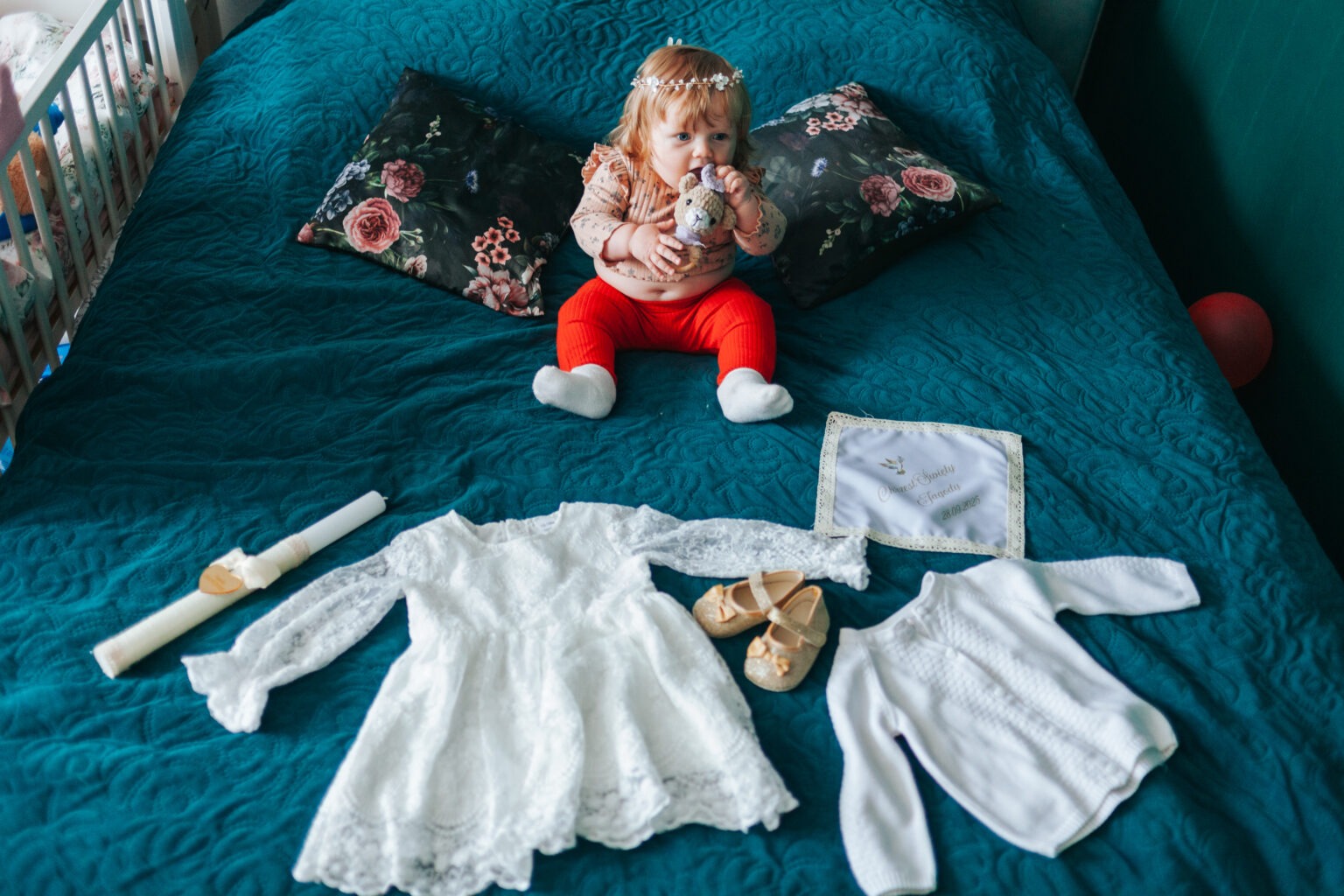 Dziewczynka z wiankiem na głowie siedzi na łóżku obok ubranka, świecy i haftowanego ręcznika z datą Chrztu Świętego. Ujęcie z góry na turkusowym tle.