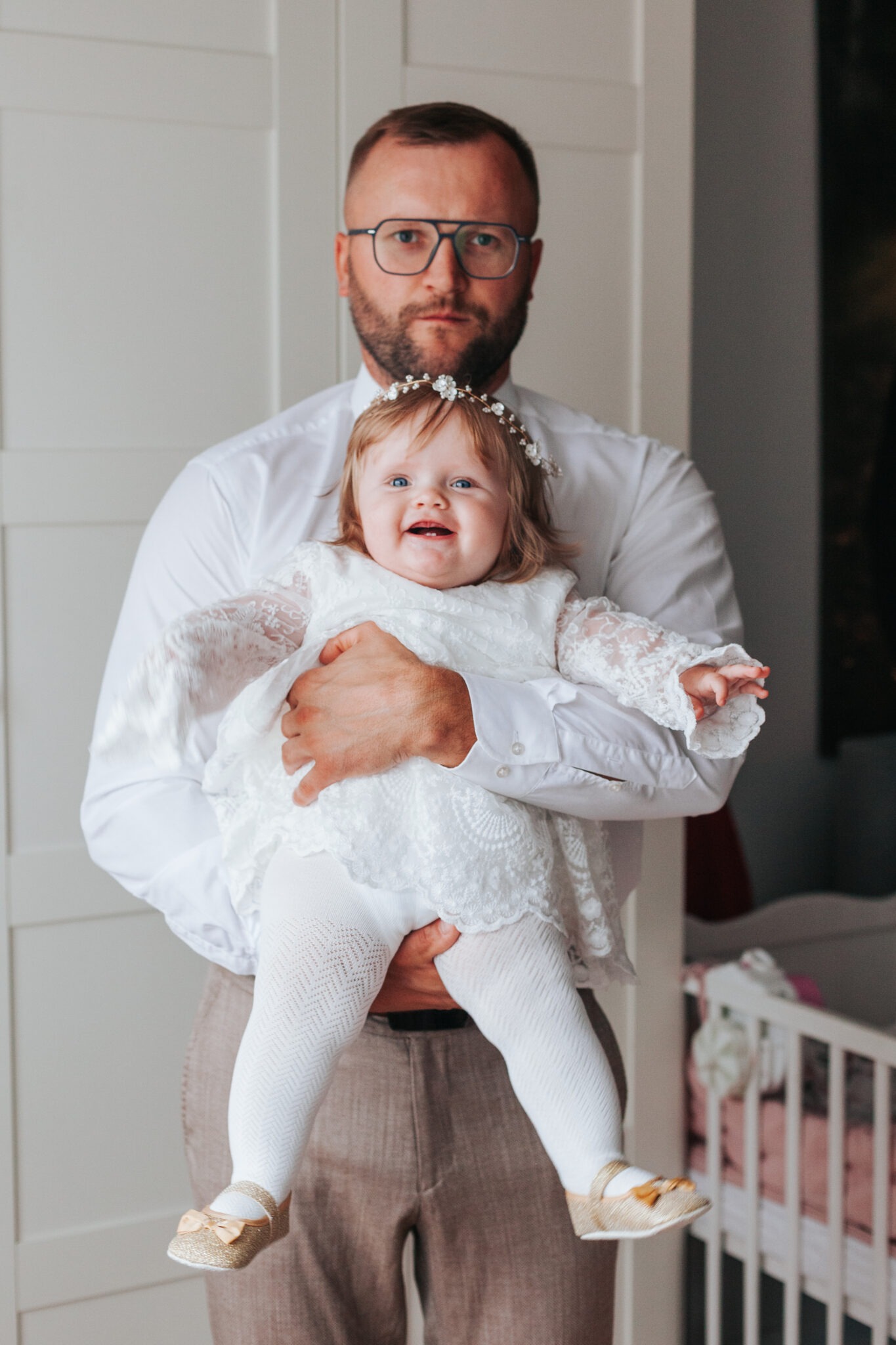 Uśmiechnięta dziewczynka w białej sukience i wianku trzymana przez mężczyznę w okularach na tle białych drzwi i łóżeczka dziecięcego.