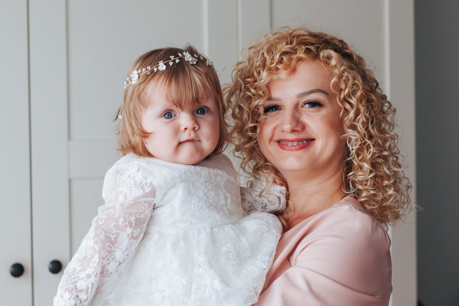 Uśmiechnięta kobieta z kręconymi włosami trzyma na rękach niemowlę w białej sukience z koronką i ozdobą na głowie, na tle białych drzwi.