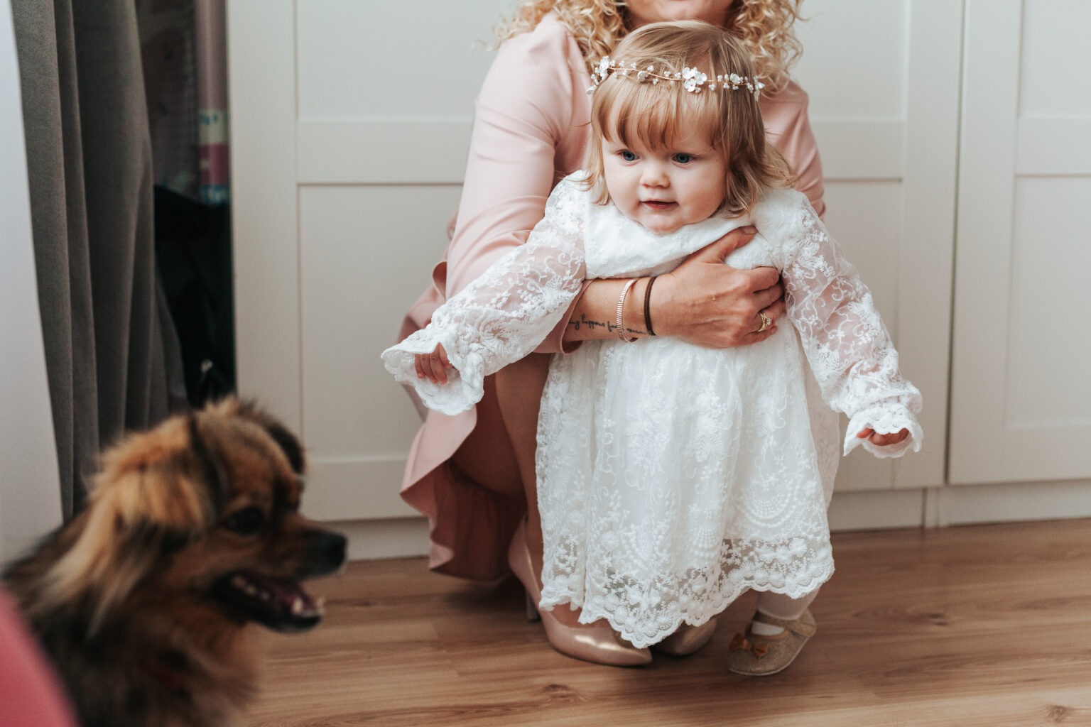 Dziewczynka w białej koronkowej sukience i wianku na głowie, trzymana przez kobietę. W tle białe meble i pies z otwartym pyskiem. Jasne, naturalne światło.