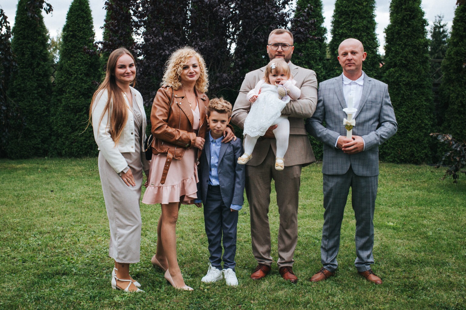 Uroczyste zdjęcie rodzinne na tle zielonego trawnika i ciemnych drzew iglastych. Mężczyzna trzyma dziecko w białej sukience, obok stoi chłopiec w garniturze i dwie kobiety.