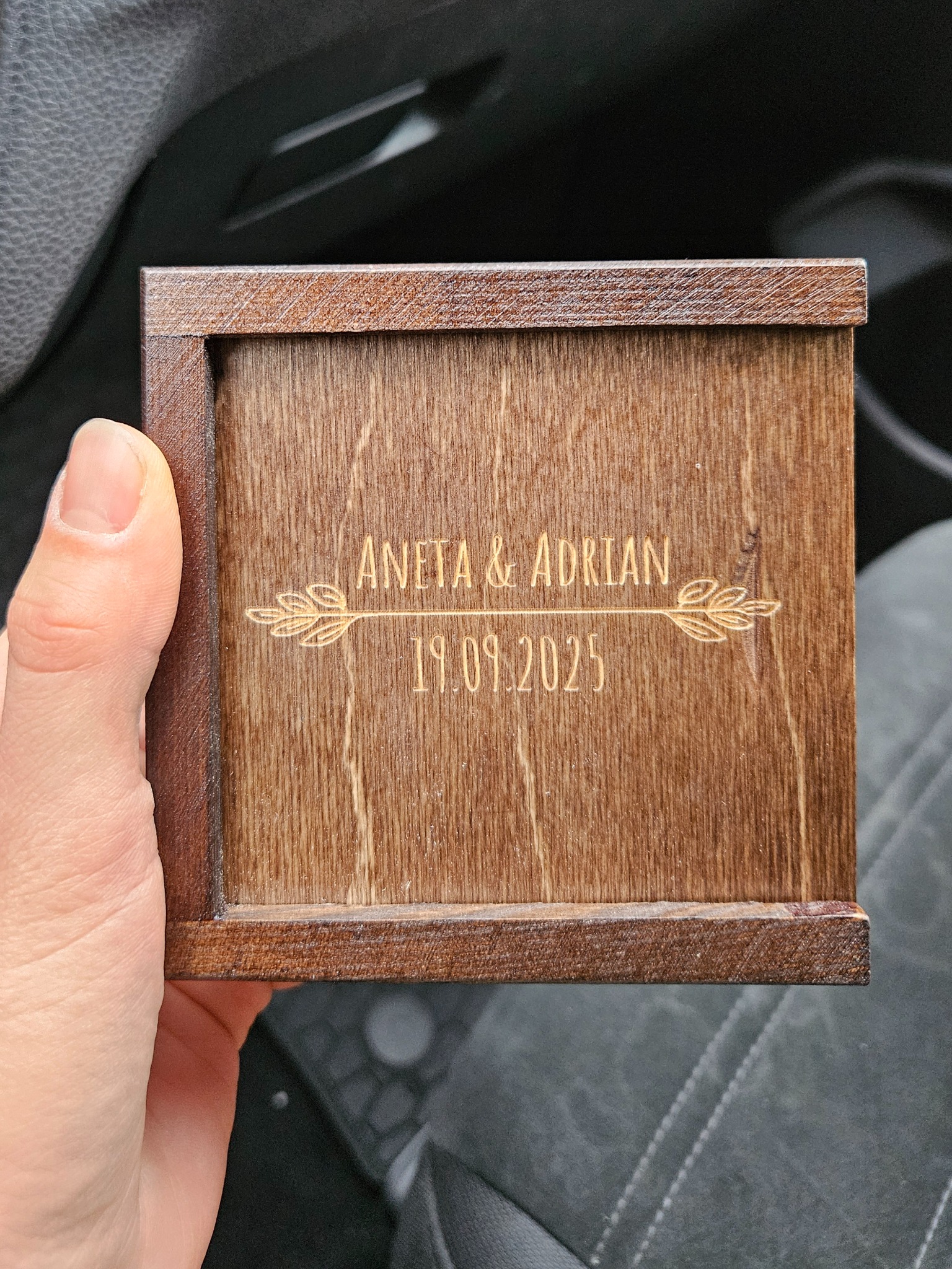 Drewniane pudełko z wygrawerowanymi imionami Aneta & Adrian oraz datą 19.09.2025, trzymane w dłoni, prawdopodobnie jako pamiątka ślubna lub prezent.