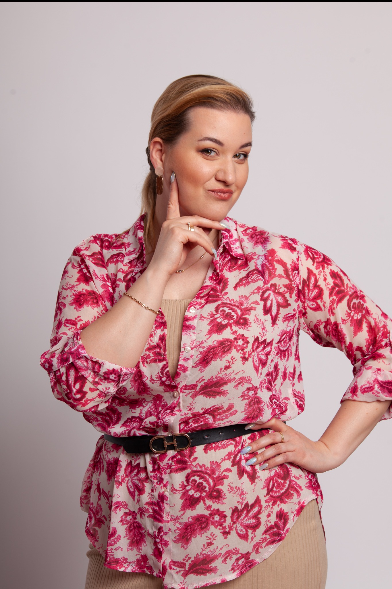 Portret kobiety w kwiecistej koszuli, z ręką pod brodą i uśmiechem, na jasnym tle. Widoczny złoty pierścionek i bransoletka, czarny pasek w talii.