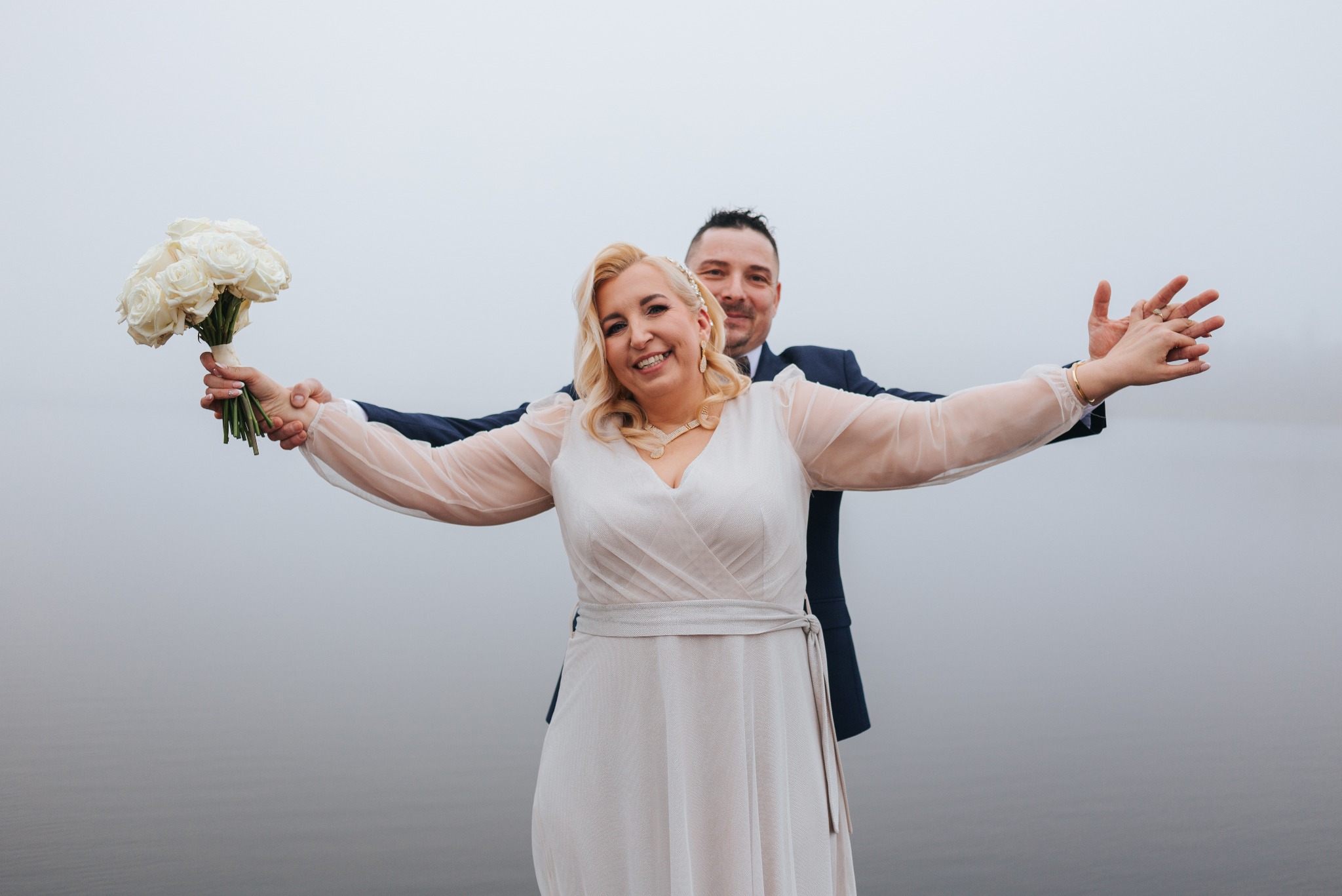 Uśmiechnięta para młoda w objęciach, ona trzyma bukiet białych róż, on obejmuje ją od tyłu. Mglista sceneria, woda w tle. Radosne uniesienie.